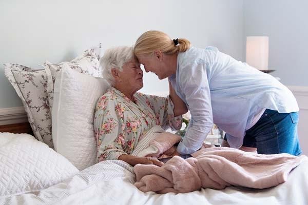 A woman leaning over an elderly woman sitting in bed, touching foreheads in a caring gesture. Caregiving at home around indianapolis
