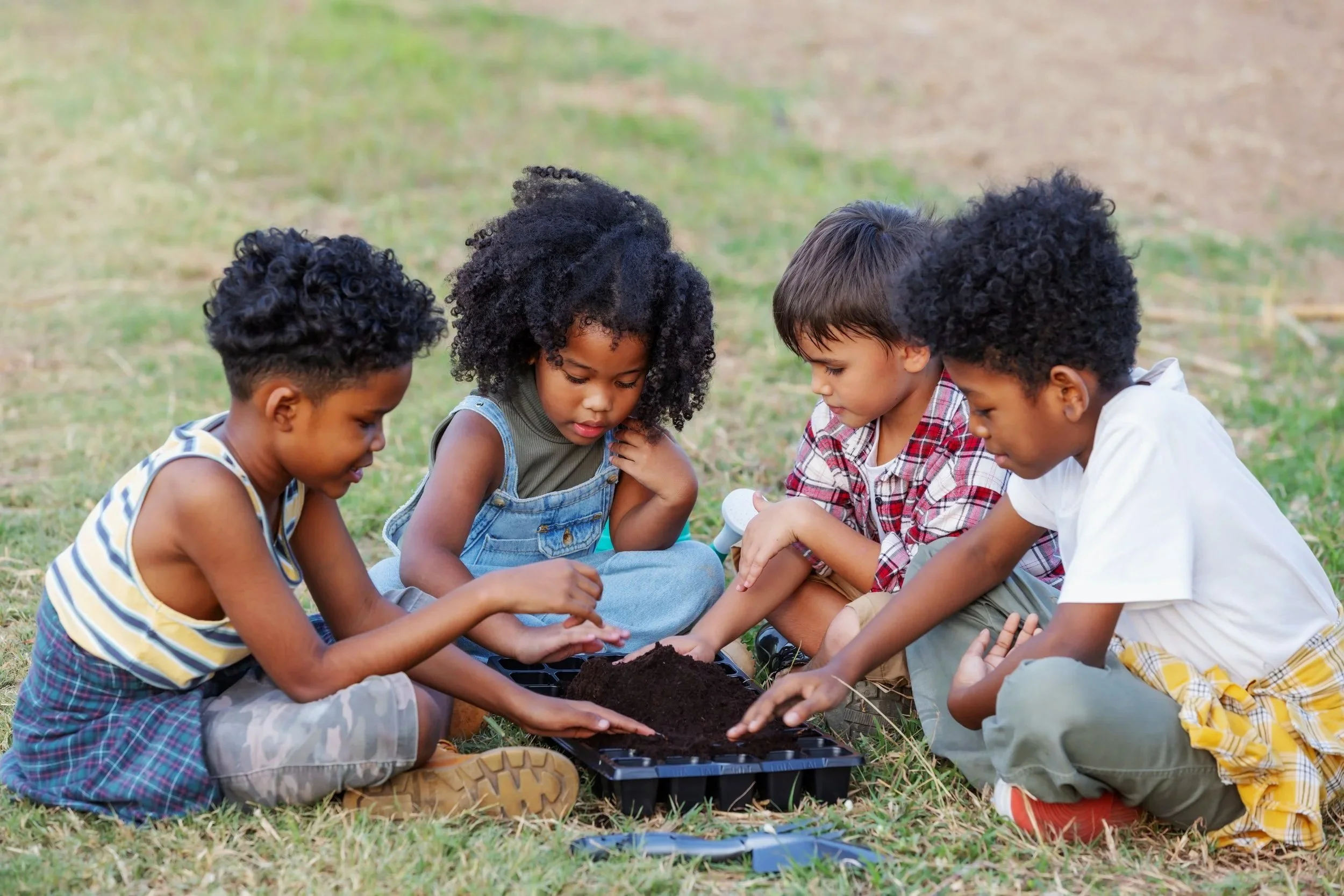 Five children sitting outdoors on grass, planting seeds or seedlings in soil, engaged in gardening activity.