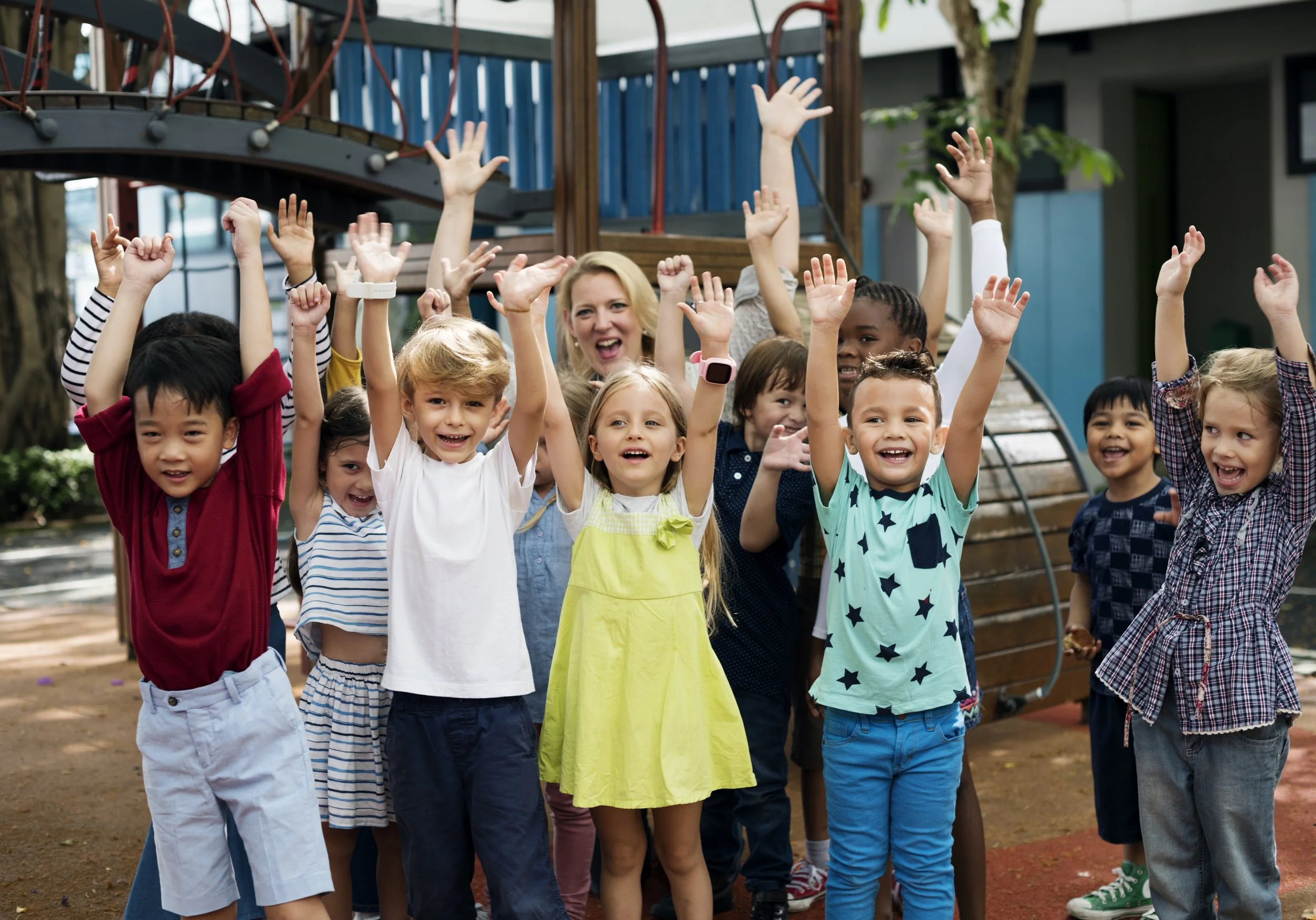 Group of children and an adult woman outdoors at a playground, raising their hands and smiling.