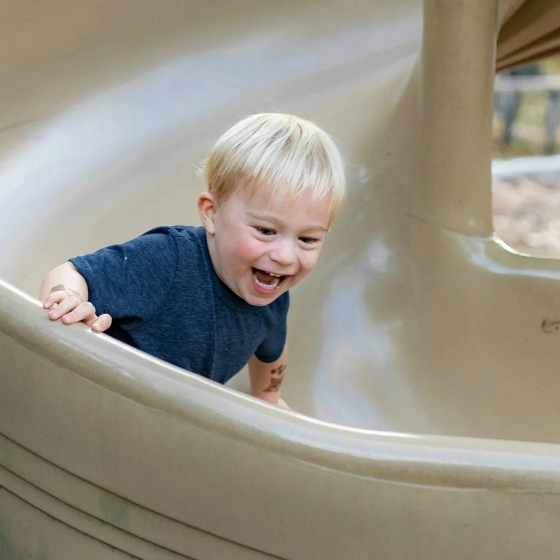 A young boy with blond hair and a blue shirt having fun on a playground slide.
