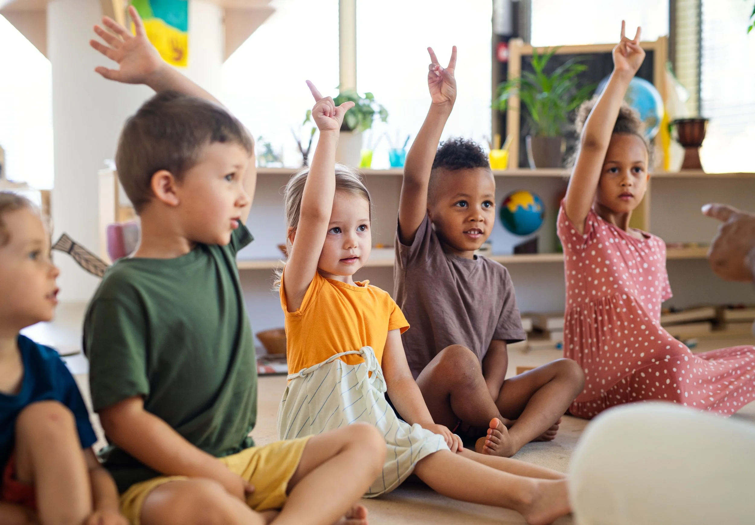 Children sitting cross-legged on the floor in a classroom, raising their hands, with some appearing eager and attentive.