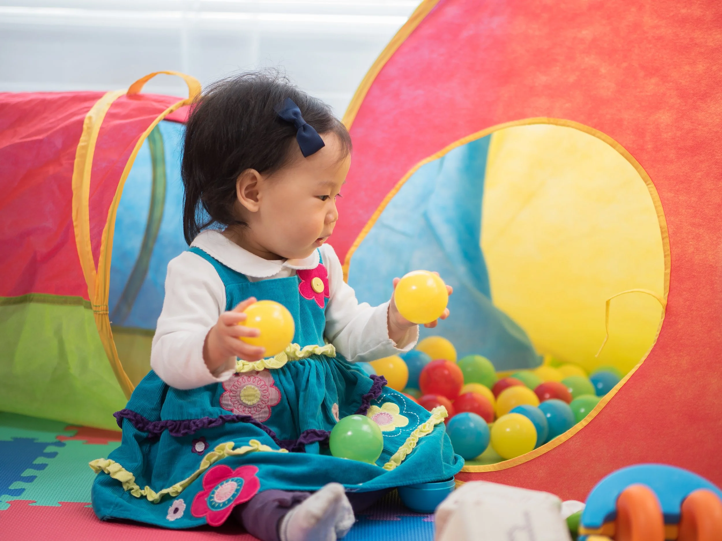 A young girl with dark hair and a navy bow, wearing a colorful dress, is sitting in a ball pit surrounded by multicolored plastic balls, with a colorful play structure in the background.