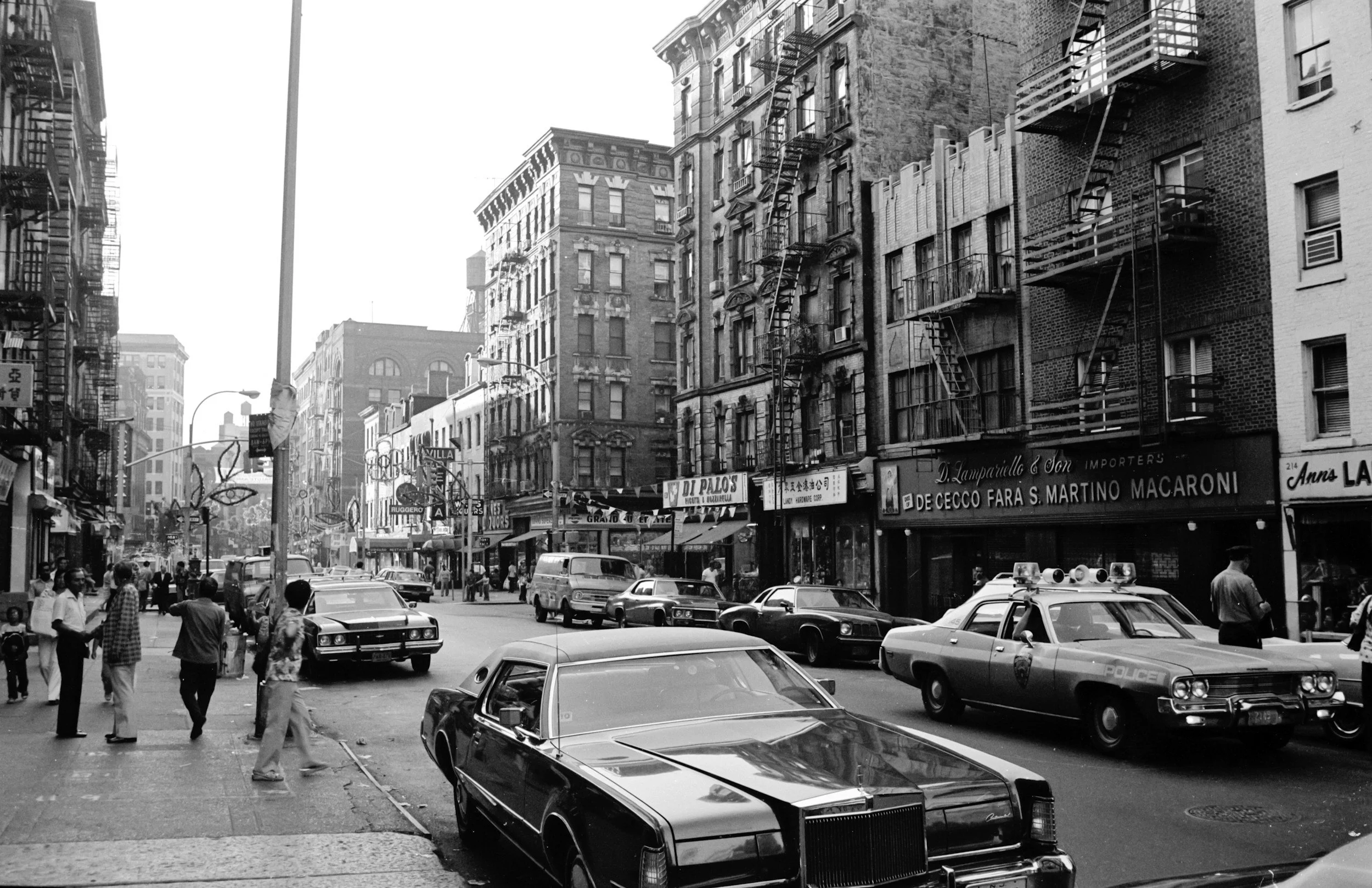 Black and white street scene with cars parked along the curb, people walking on the sidewalk, and buildings with fire escapes and storefronts.