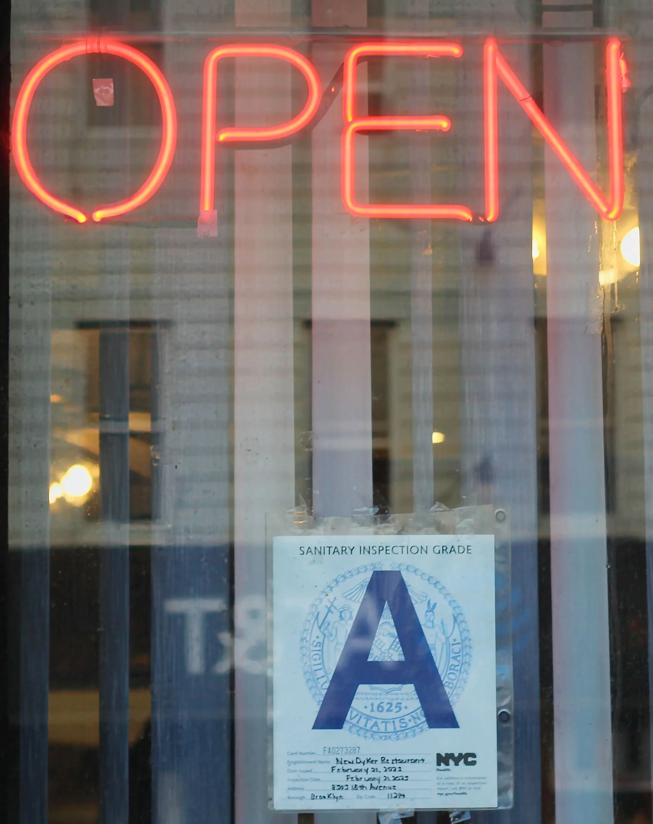 A neon red 'OPEN' sign hanging in a window with a Sanitary Inspection Grade 'A' certificate from NYC posted below.
