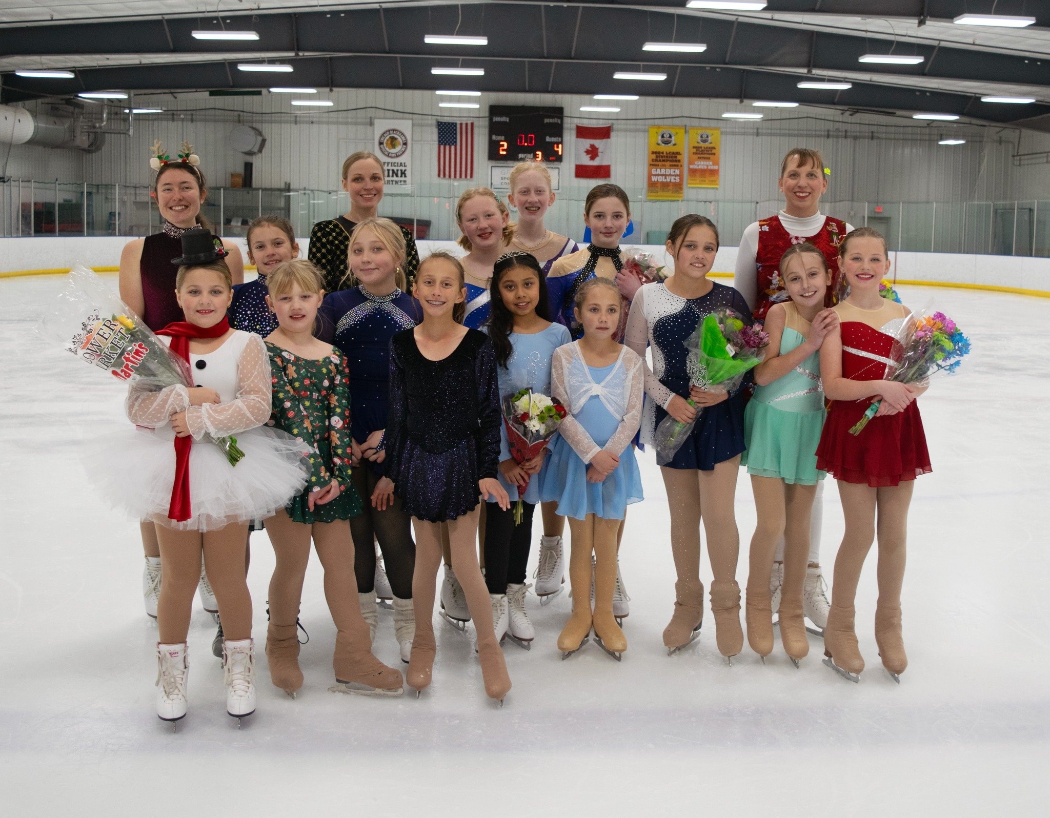 Group of young figure skaters in colorful costumes posing with coaches or instructors on an ice rink after a performance or competition, some holding flowers.