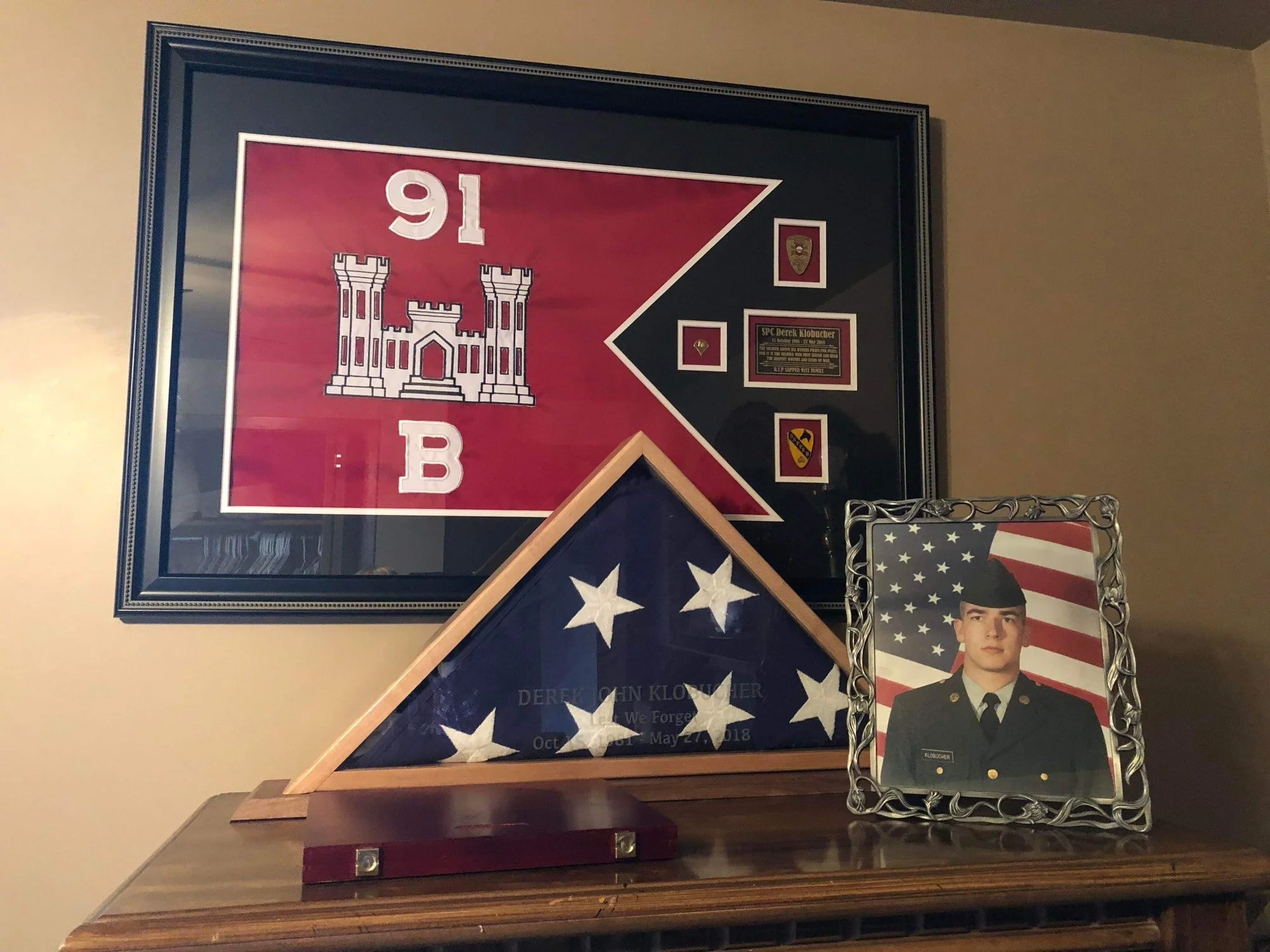 A framed red and black flag with a white castle emblem, military patches, and a plaque. A wooden display case with a folded American flag and text honoring Derek John Klobucher. A framed photo of a young soldier in uniform with an American flag background.