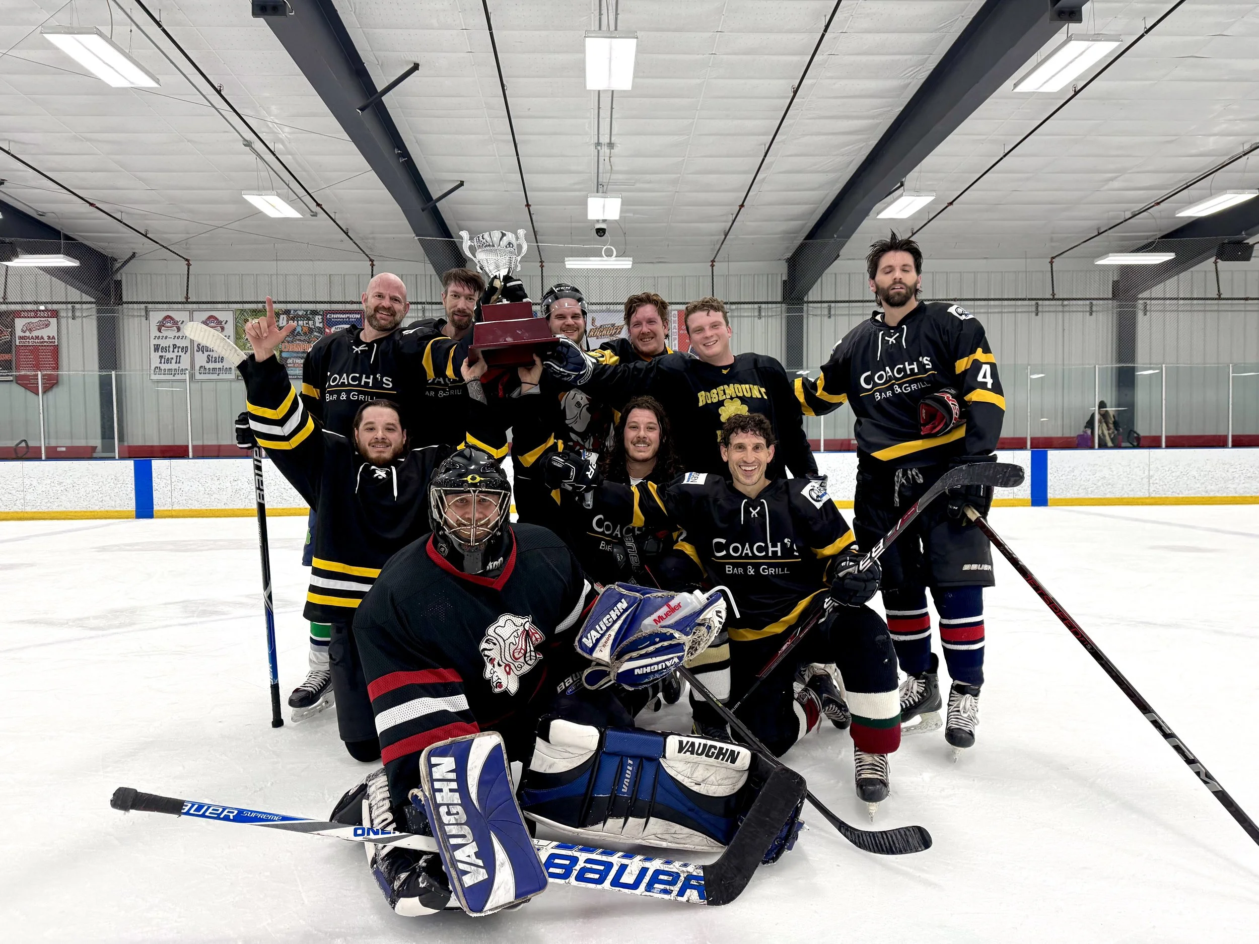 A group of ice hockey players celebrating on the ice rink, holding a trophy. They are wearing black jerseys with yellow stripes, and there's a goalie in front wearing a black jersey with a lion logo. The team members are smiling and posing for the photo after a game.
