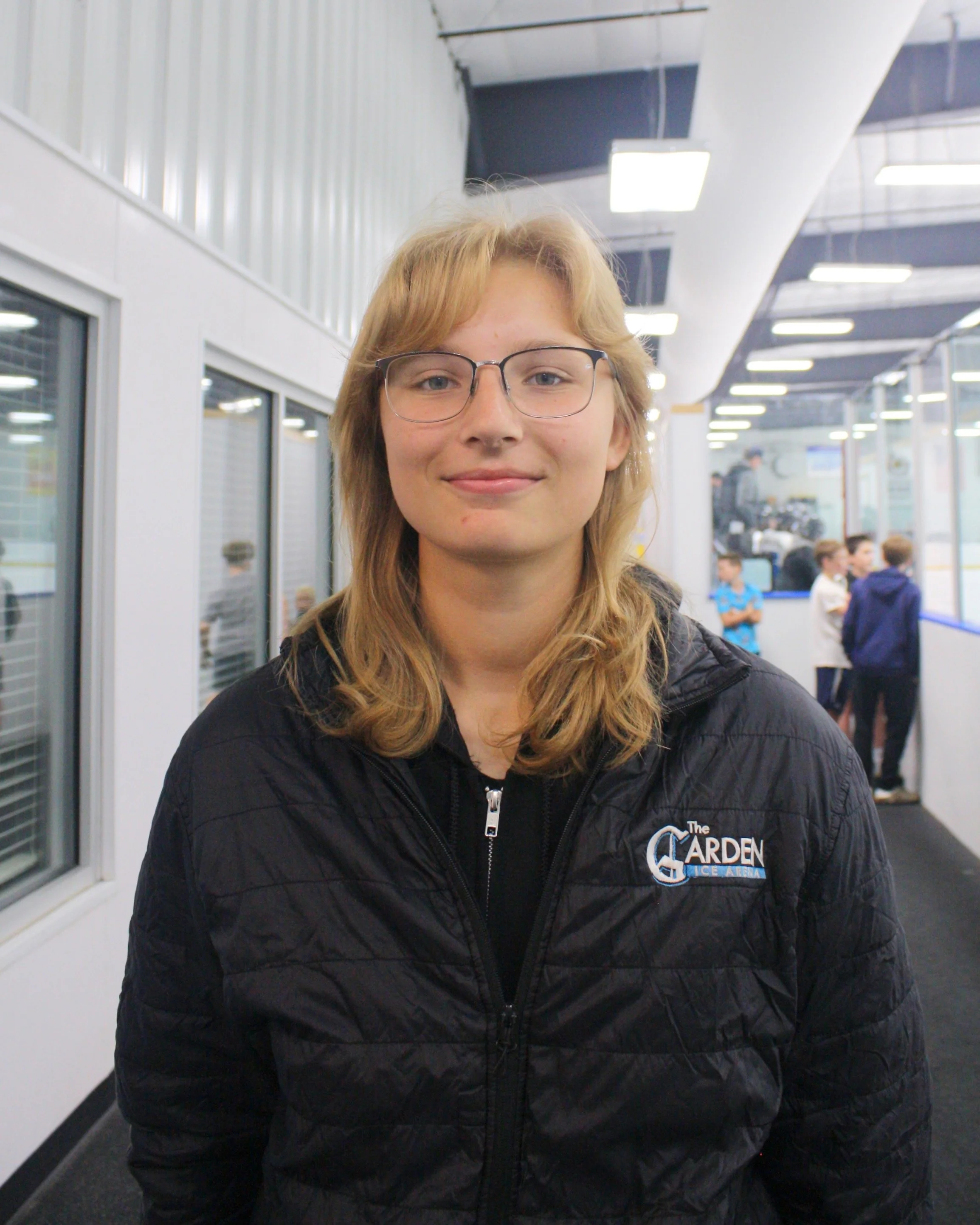 A young woman with blonde hair wearing a black quilted jacket with 'The Garden Ice Arena' logo, standing indoors with a modern rink background.