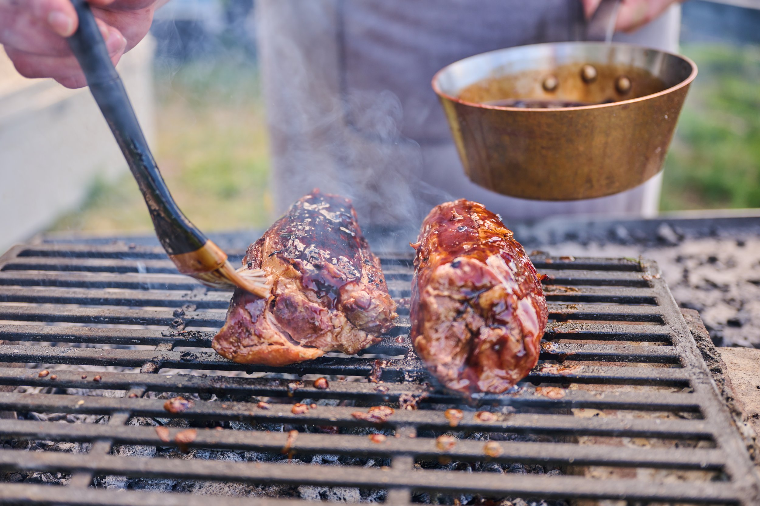 Two large pieces of meat are being grilled on a barbecue grill, with a person using tongs to handle one piece. Smoke rises from the meat, which is glazed with barbecue sauce.