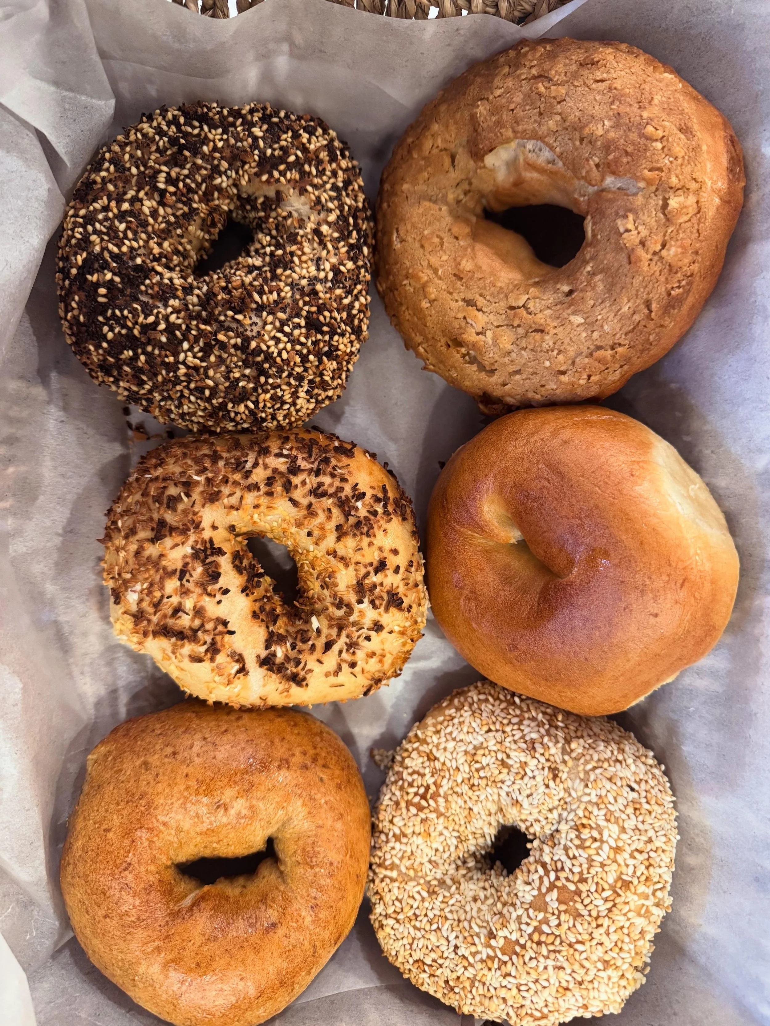 Six assorted bagels on a white paper-lined tray, including ones with sesame seeds, chocolate chips, chopped nuts, and a plain bagel.