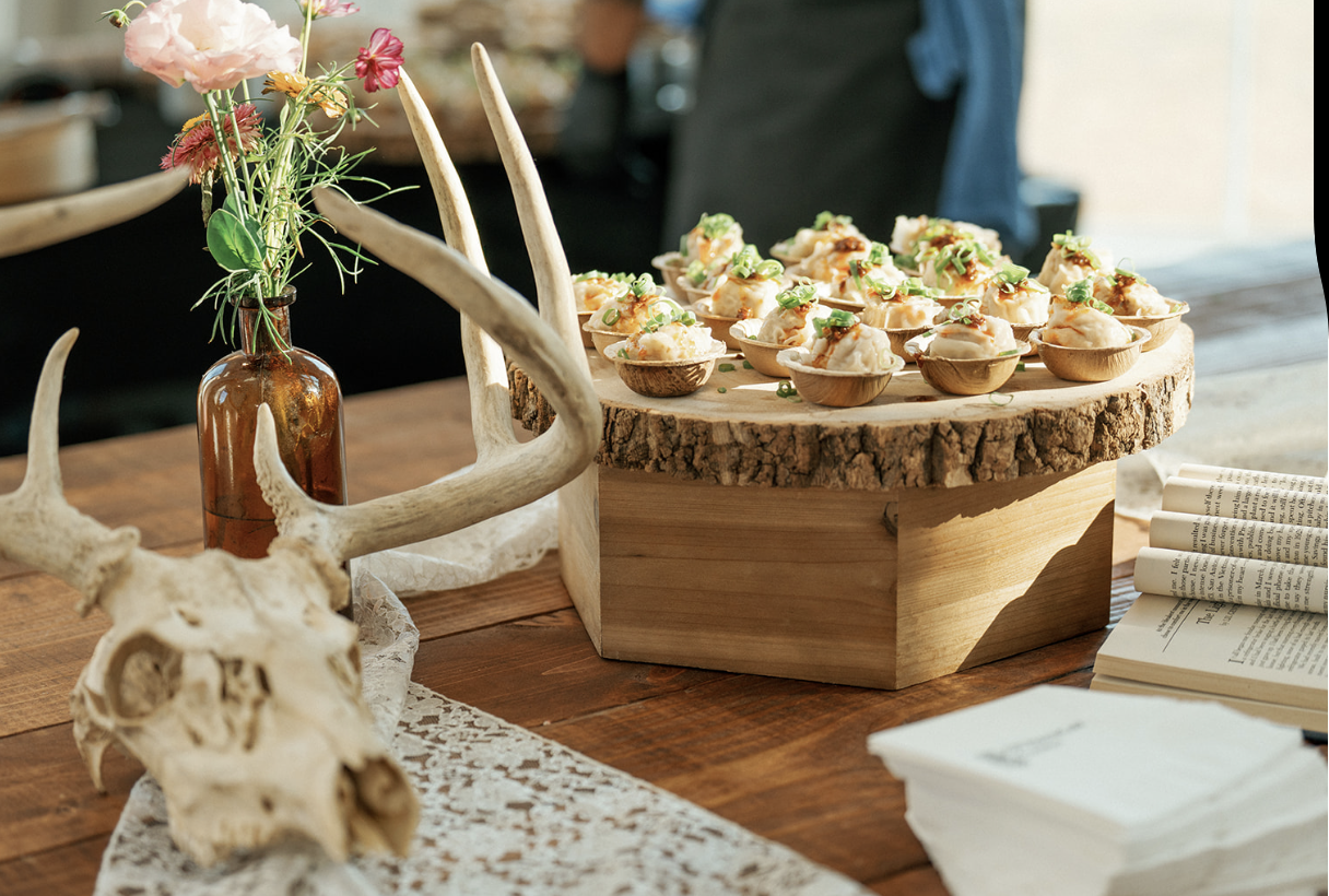 A wooden table with a centerpiece of an antler and a brown glass vase holding pink and purple flowers. Next to the centerpiece, there is a large wooden slab with small topped snacks or appetizers garnished with green onions. To the right, there are r
