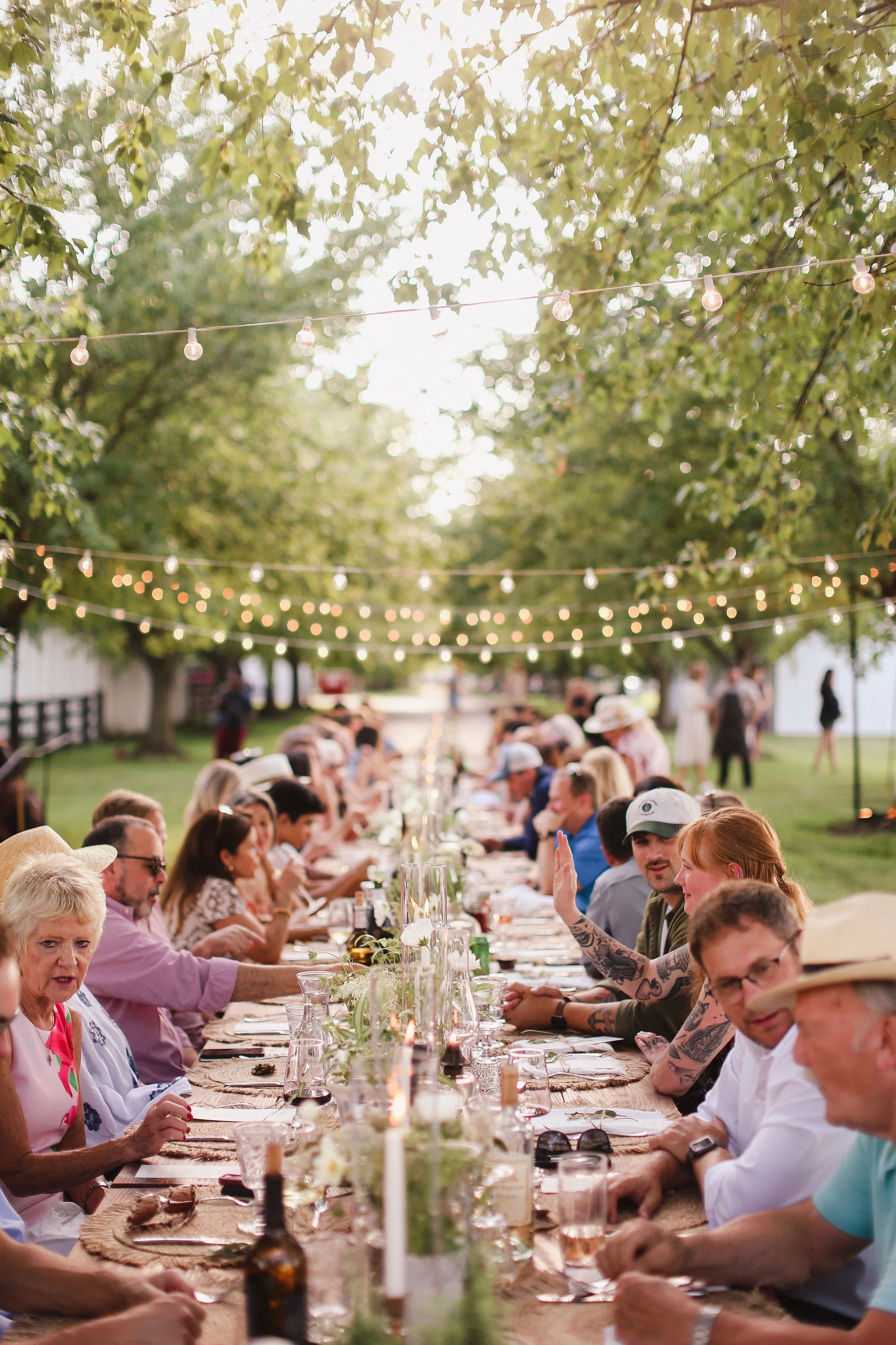 People are gathered around a long outdoor dining table beneath string lights, enjoying a social event in a green outdoor setting.