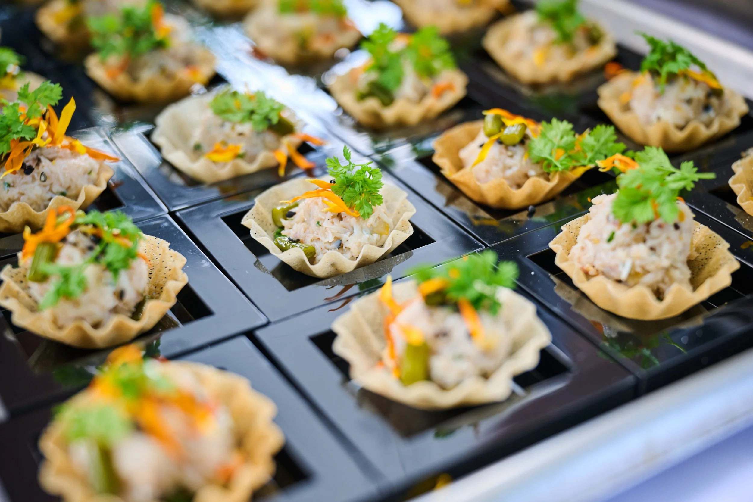 Miniature savory tarts filled with a mixture of seafood or chicken salad, topped with fresh cilantro, and garnished with orange and green vegetables.