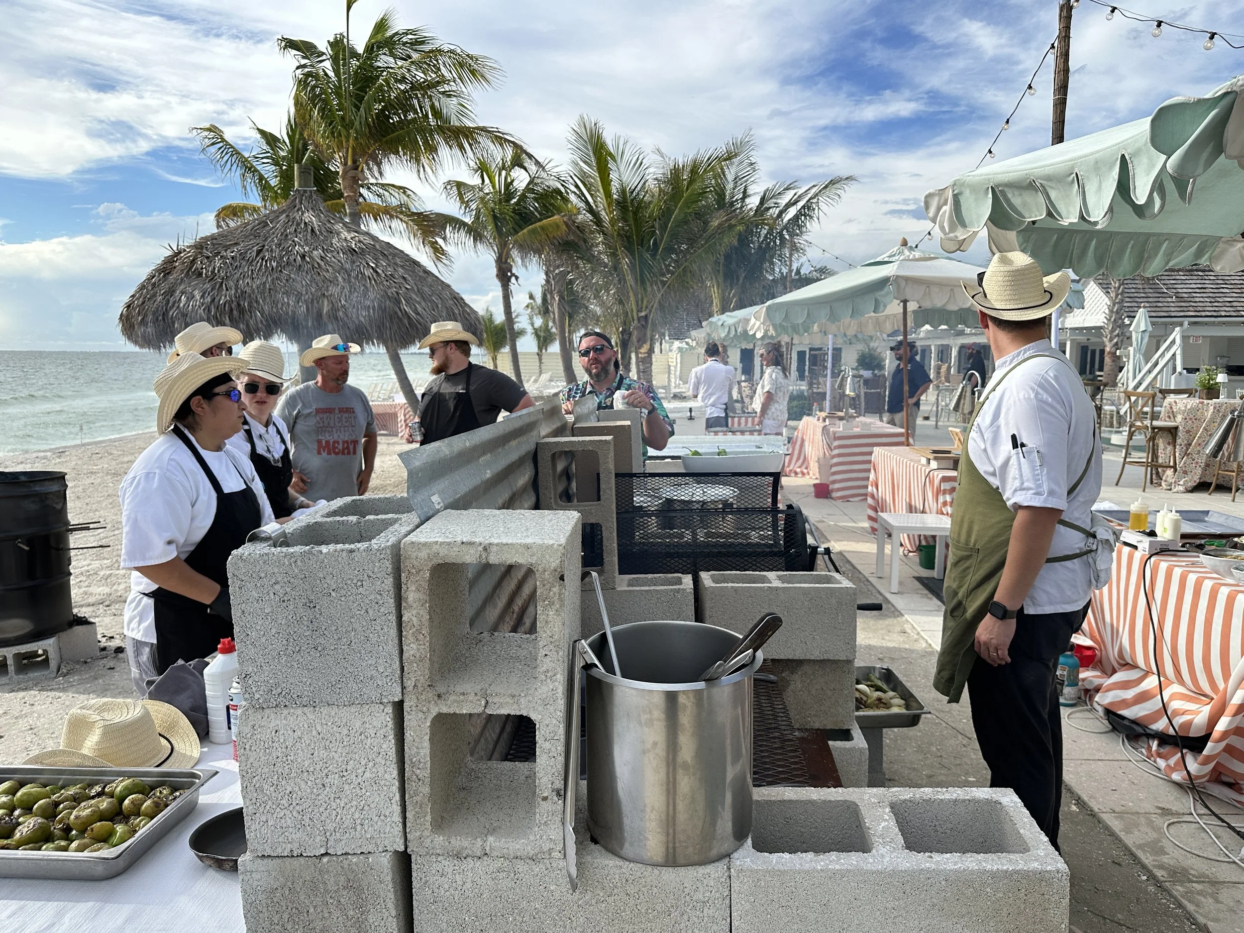 Chef Josh and staff cooking seafood on an outdoor grill by the beach with palm trees, umbrellas, and tables set for dining.