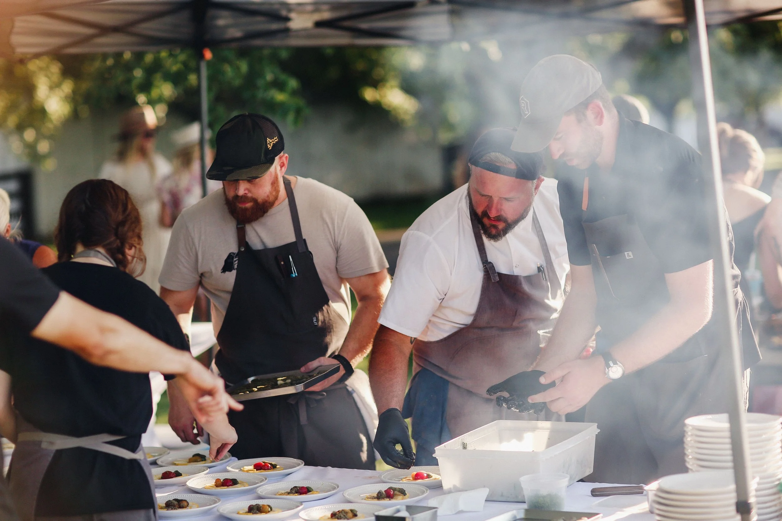 Chef Josh plating food outdoors at a food event under a canopy, with plates of food arranged on a table.