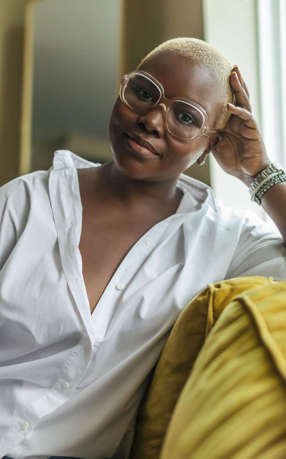 A woman with short blond hair wearing large clear glasses and a white shirt, sitting on a yellow couch, resting her head on her hand, looking at the camera, with a calm expression.