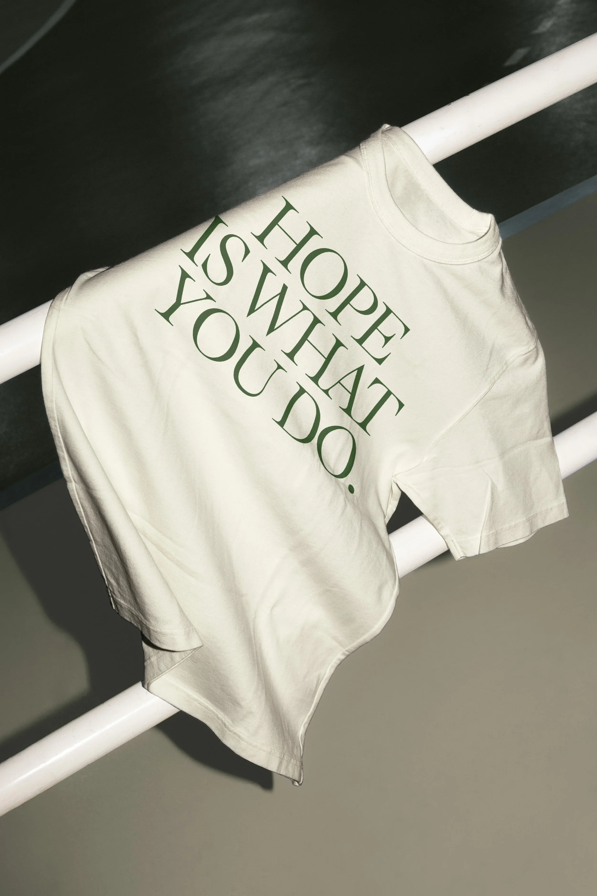 A white T-shirt hanging on a white metal rod with the phrase 'HOPE IS WISH YOU DO.' written in large green letters.