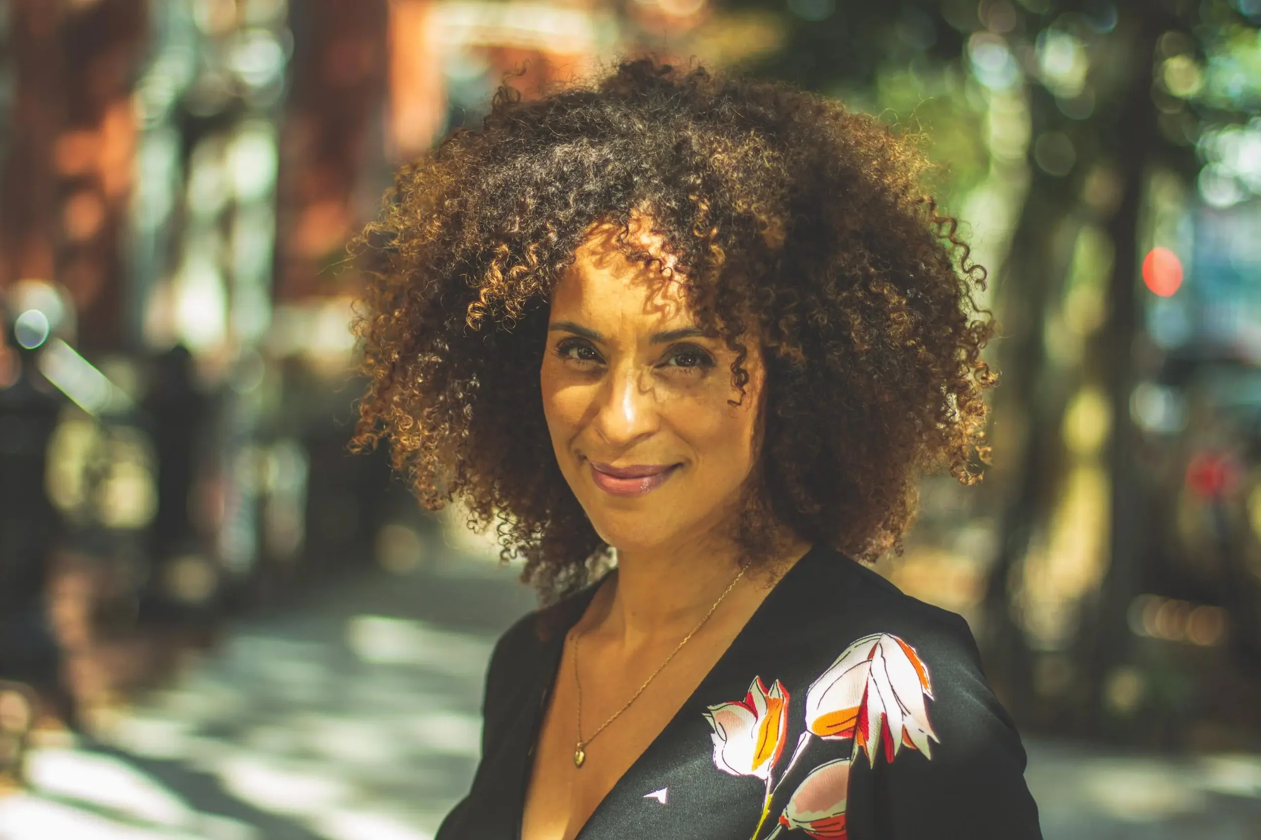 A woman with curly brown hair smiling outdoors on a sunny day, wearing a black top with floral pattern, a gold necklace, and a necklace with a small pendant.