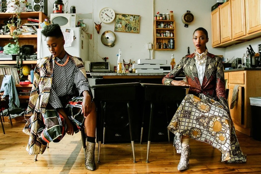 Two women in fashionable outfits sitting in a cozy, cluttered kitchen with wooden cabinets, spices, and kitchen appliances on shelves and counters.