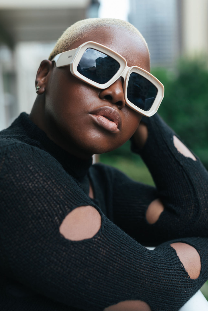 A woman with short platinum blonde hair wearing large white sunglasses and a black ripped sweater, outdoors with urban buildings in the background.