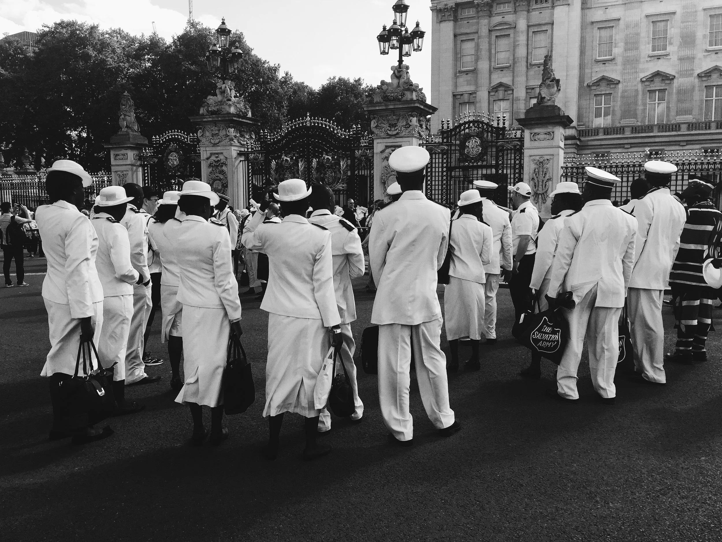 A group of people in white uniforms, some wearing hats, gathered outside a gated area with ornate gates and a large building in the background.