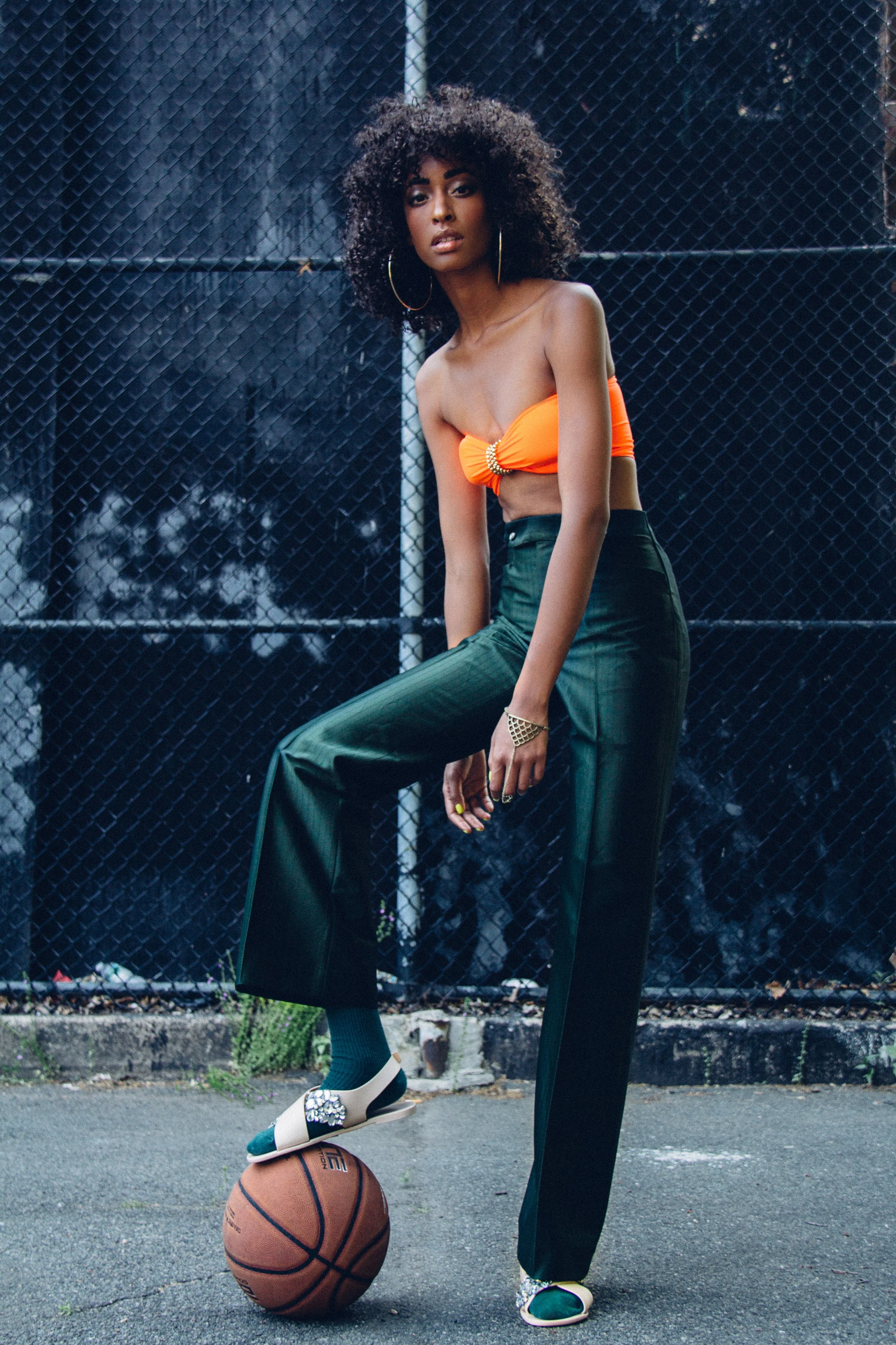 A woman with an afro hairstyle, wearing a bright orange strapless top, dark green high-waisted pants, and slides, standing on a basketball with one foot, on an outdoor court with a chain-link fence in the background.
