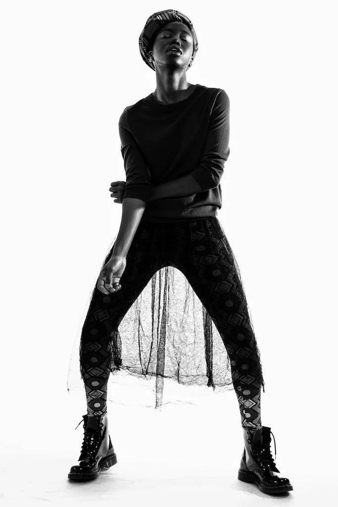 A woman wearing a beret, a dark long-sleeve top, patterned leggings, and lace-up boots poses with one hand on her leg and the other crossing her body in a studio setting with a plain background.