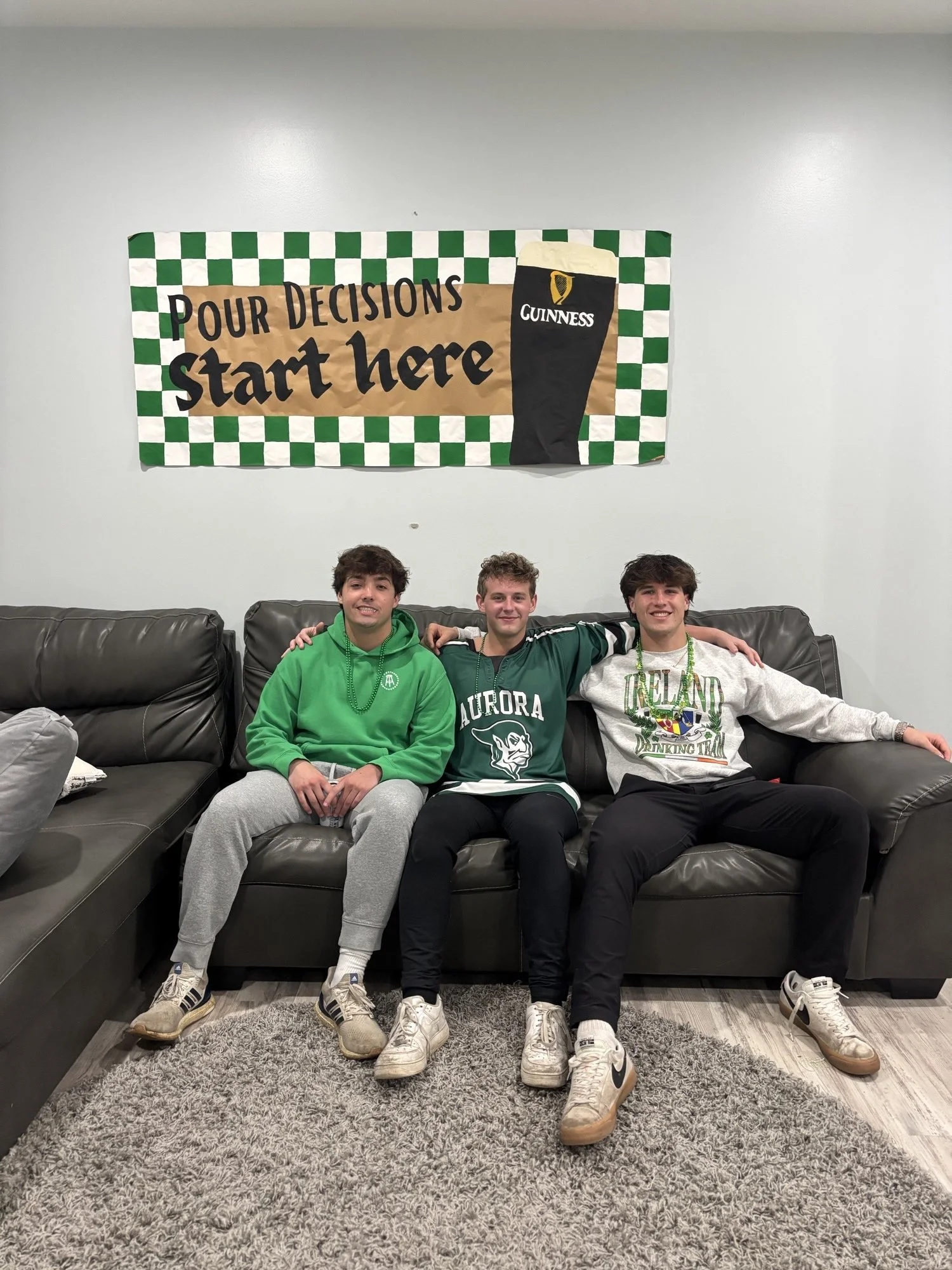 Three young men sitting on a black leather couch, smiling, with a banner behind them that reads 'Pour Decisions Start Here' and features a pint of Guinness and a checkered green and white pattern.