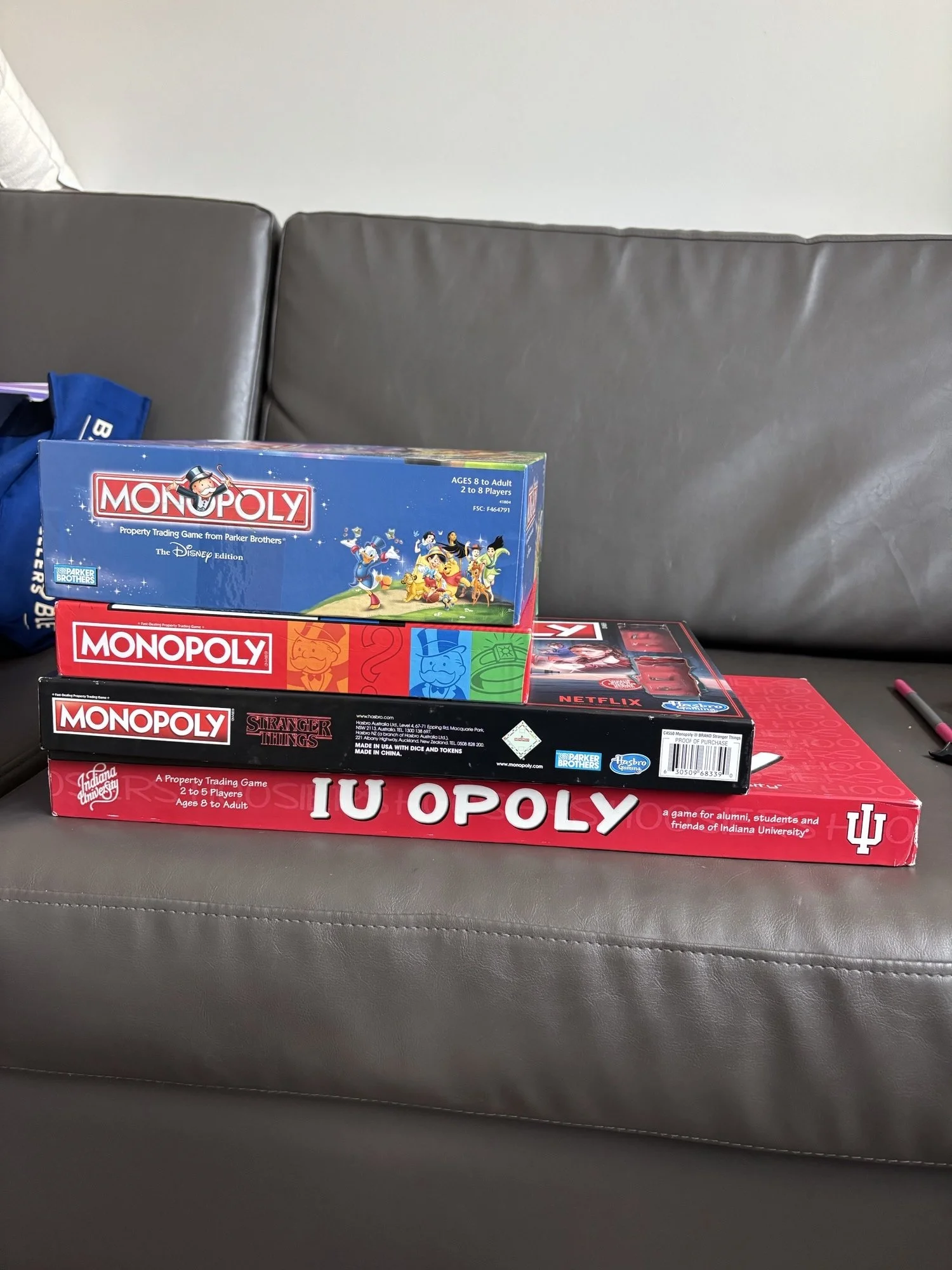 Stacked board games on a gray couch, including Monopoly Disney edition, Monopoly Junior, Stranger Things Monopoly, and IUopoly.