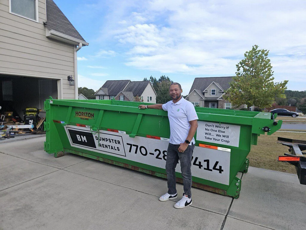 A man standing next to a large green dumpster in a residential driveway, with houses and trees in the background.