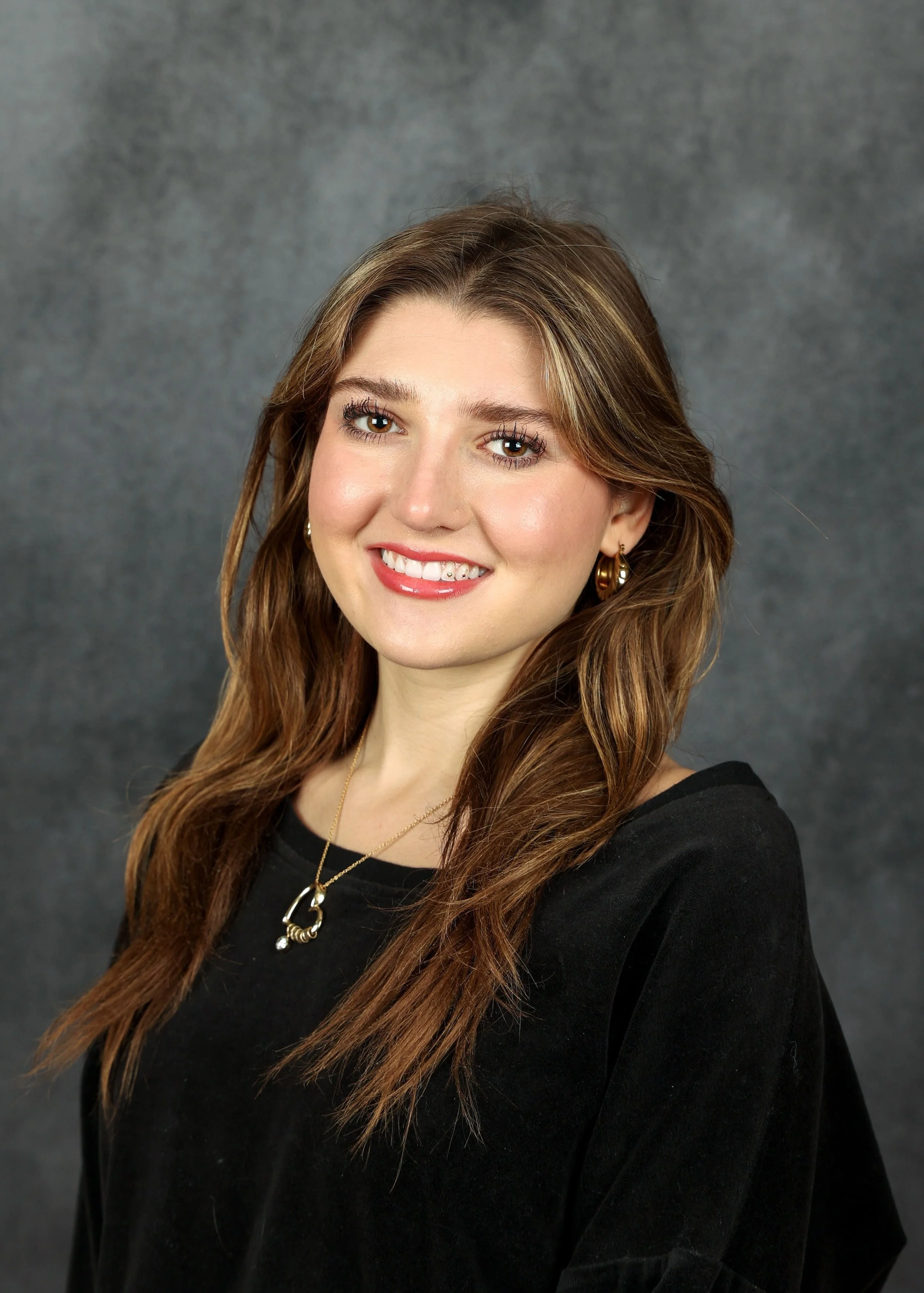 A smiling woman with long brown hair, wearing gold earrings and a gold necklace, dressed in a black top, against a gray textured background.
