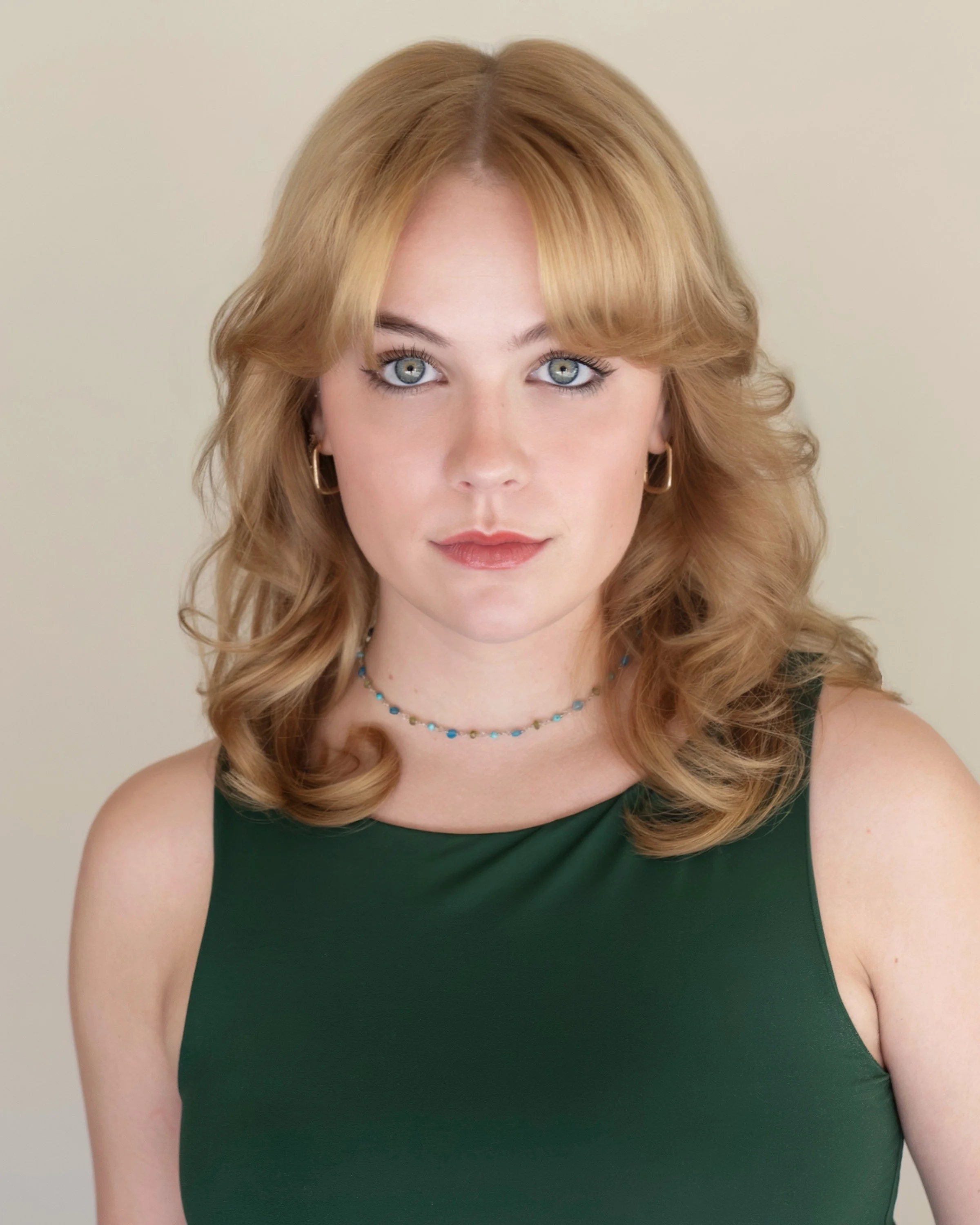 A young woman with wavy red hair, blue eyes, and light skin, wearing a dark green sleeveless top, gold hoop earrings, a blue beaded necklace, and a subtle makeup look, standing against a light-colored background.