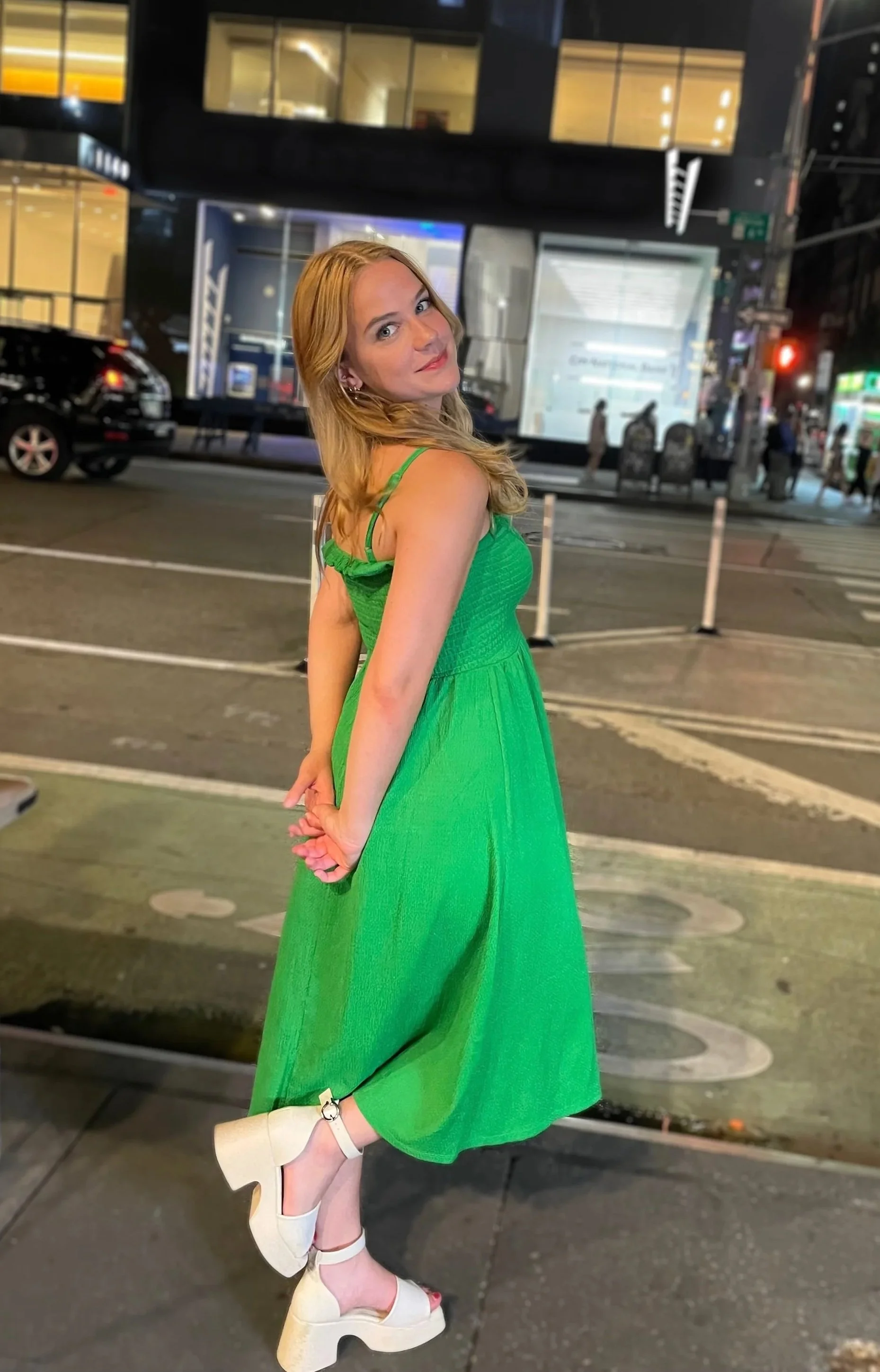 A woman in a bright green dress and white platform shoes standing on a city sidewalk at night, with buildings and cars in the background.