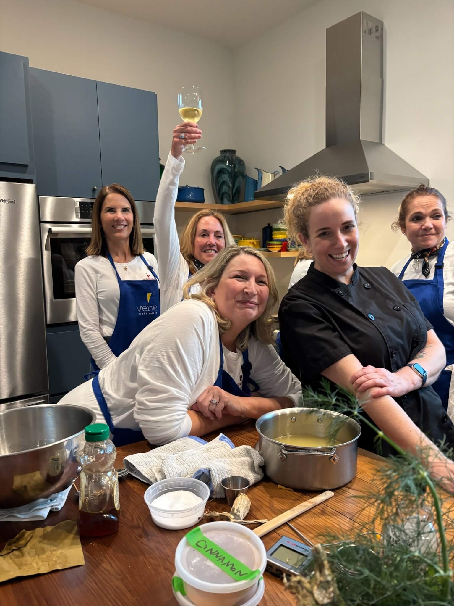 Group of five women in a kitchen, two are wearing aprons, one woman is holding a glass of white wine up in the air. The kitchen has blue cabinets and various cooking utensils and ingredients on the table.