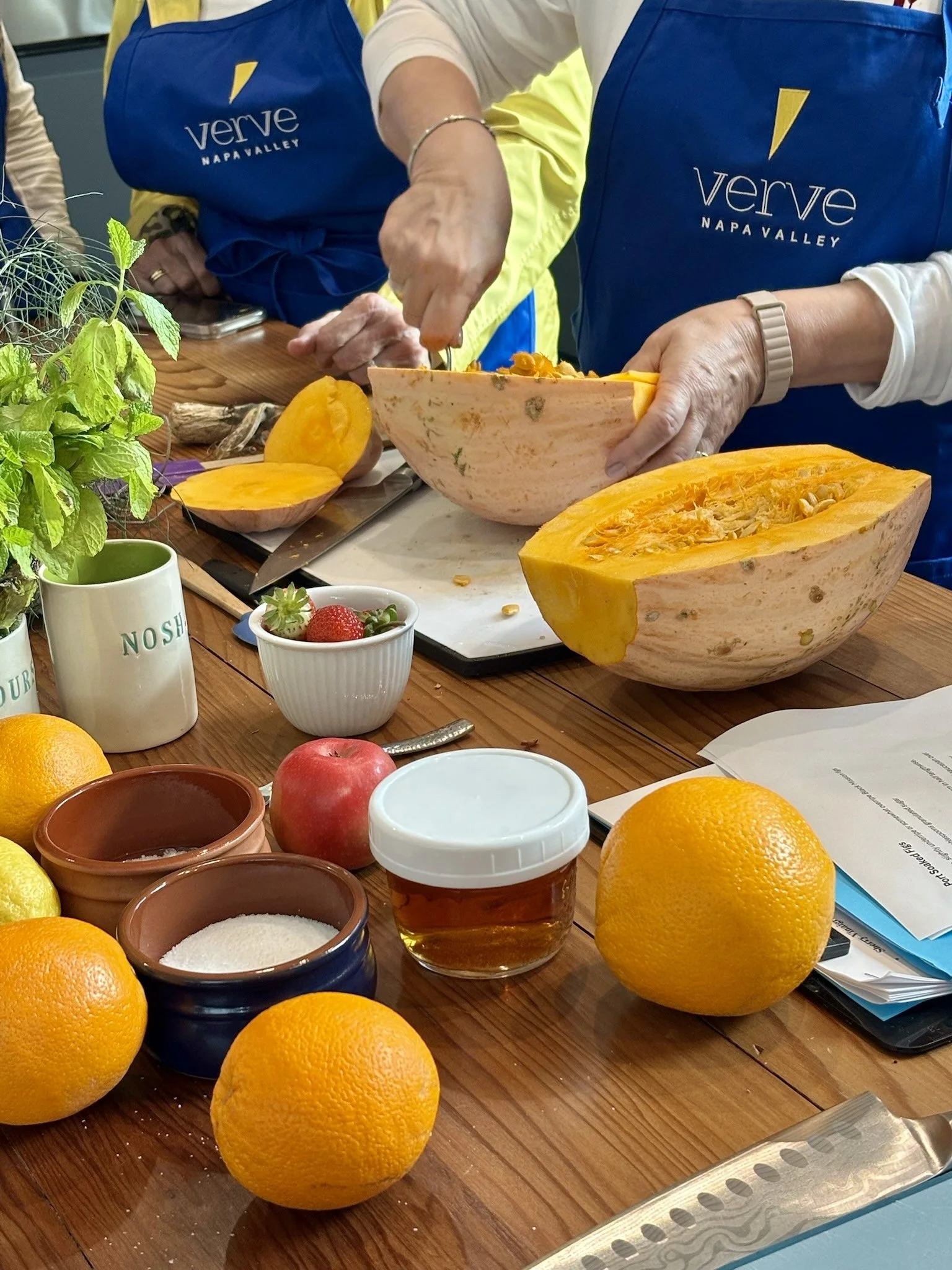 People preparing a pumpkin for cooking or carving at a kitchen table with oranges, apples, honey, salt, strawberries, and various herbs and utensils.
