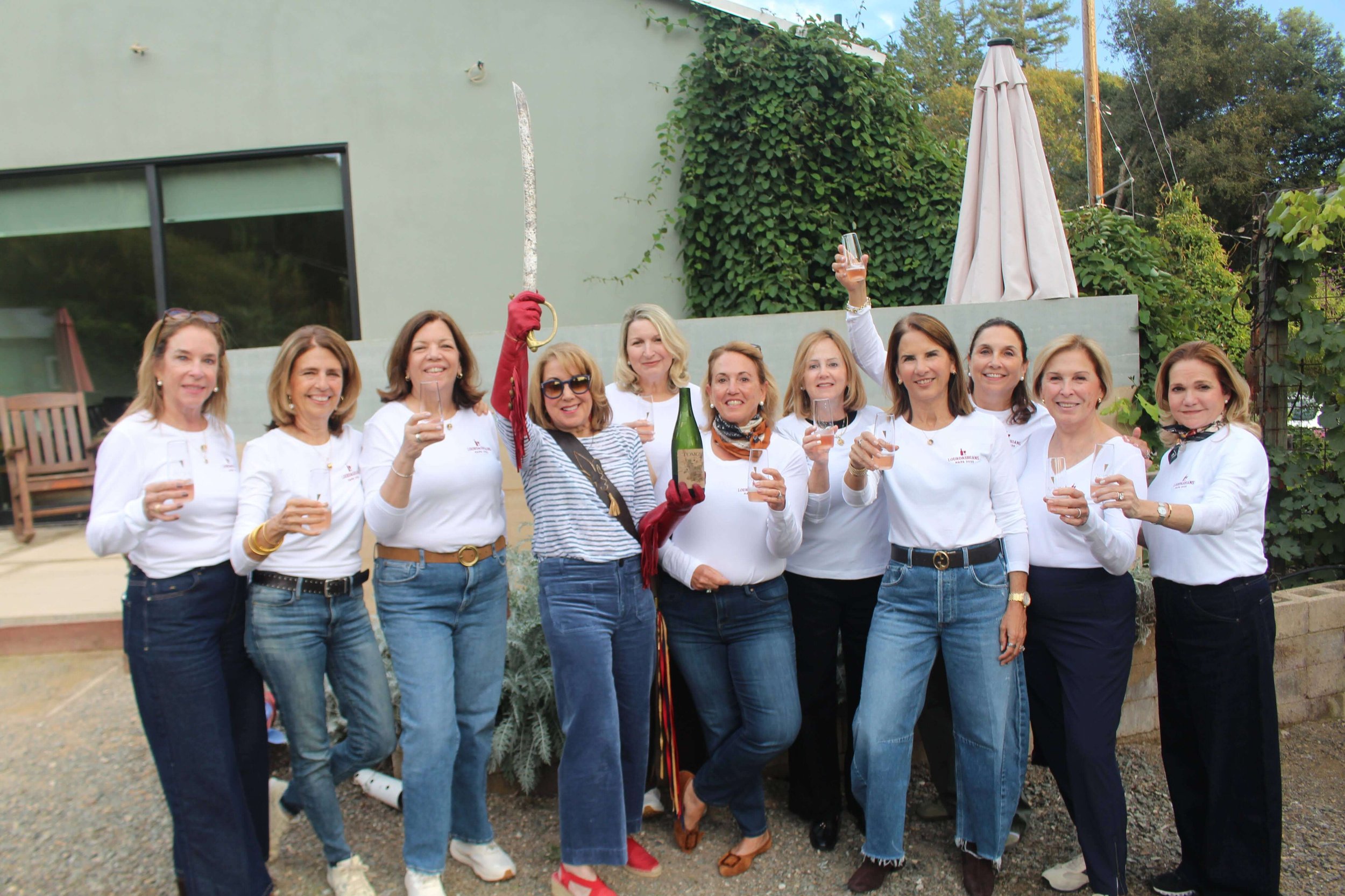 Group of eleven women at an outdoor celebration, some holding glasses of champagne, one woman in the center holding a bottle and a sword, smiling and raising glasses in a toast.