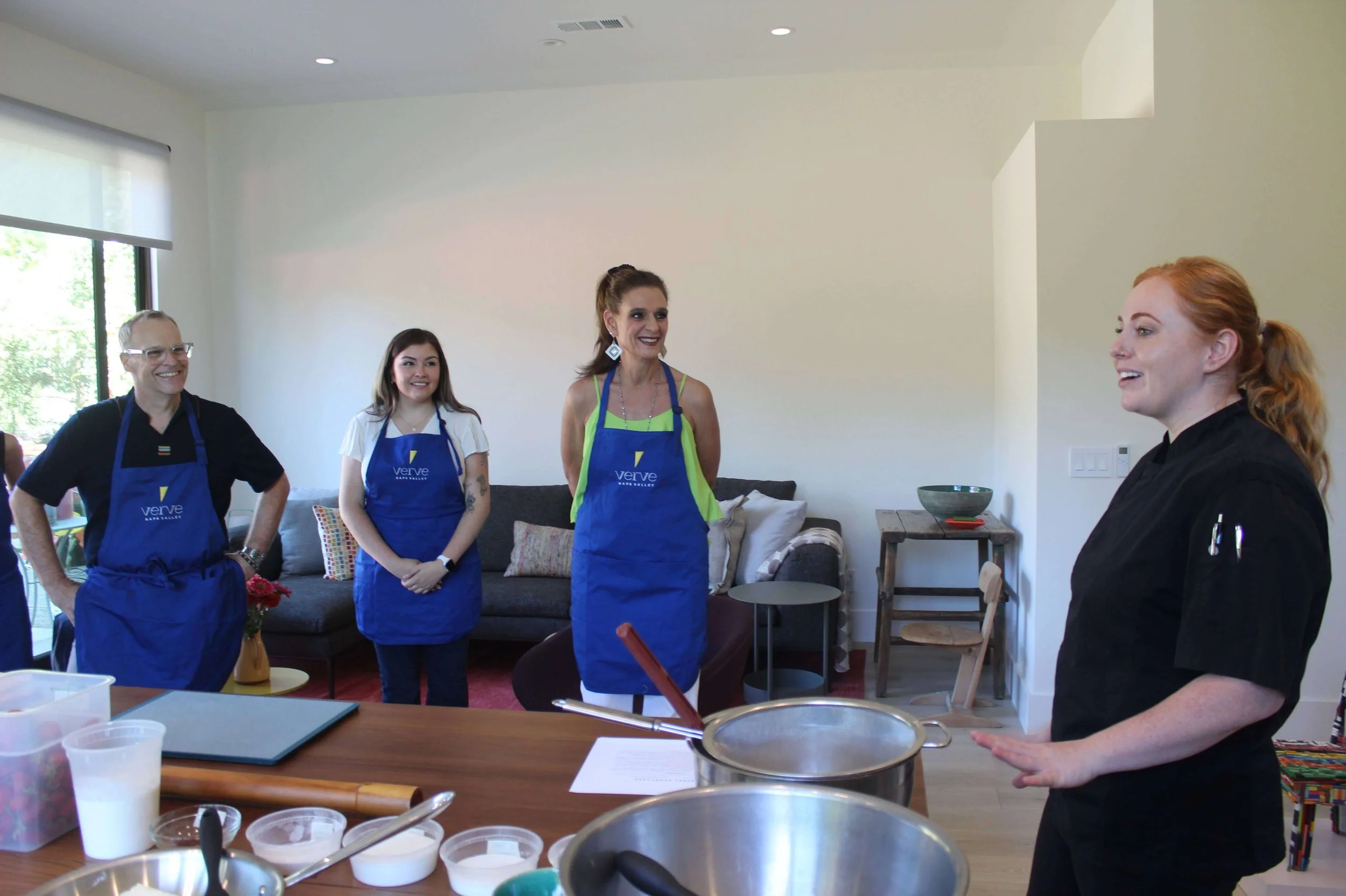 Five people in a kitchen: four women and one man, all wearing aprons, engaged in a cooking class or demonstration, with a woman speaking to the group.