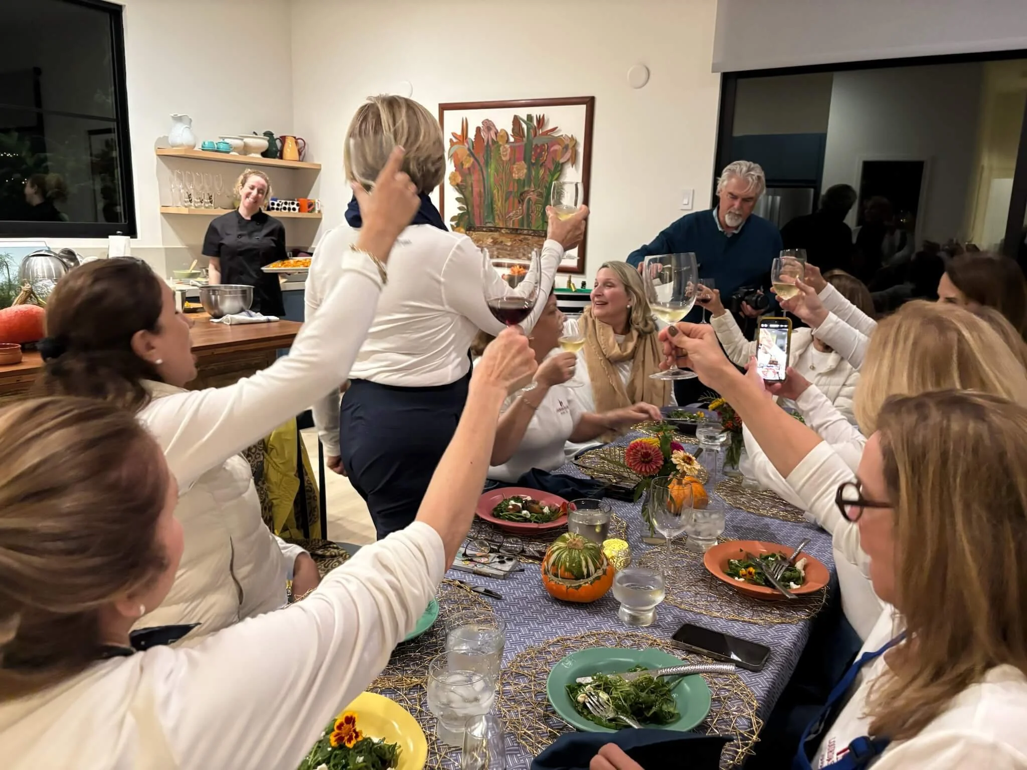 People celebrating at a dinner party, raising glasses for a toast, with a woman in the center holding a glass of white wine. The table is decorated with pumpkins, flowers, and various dishes.