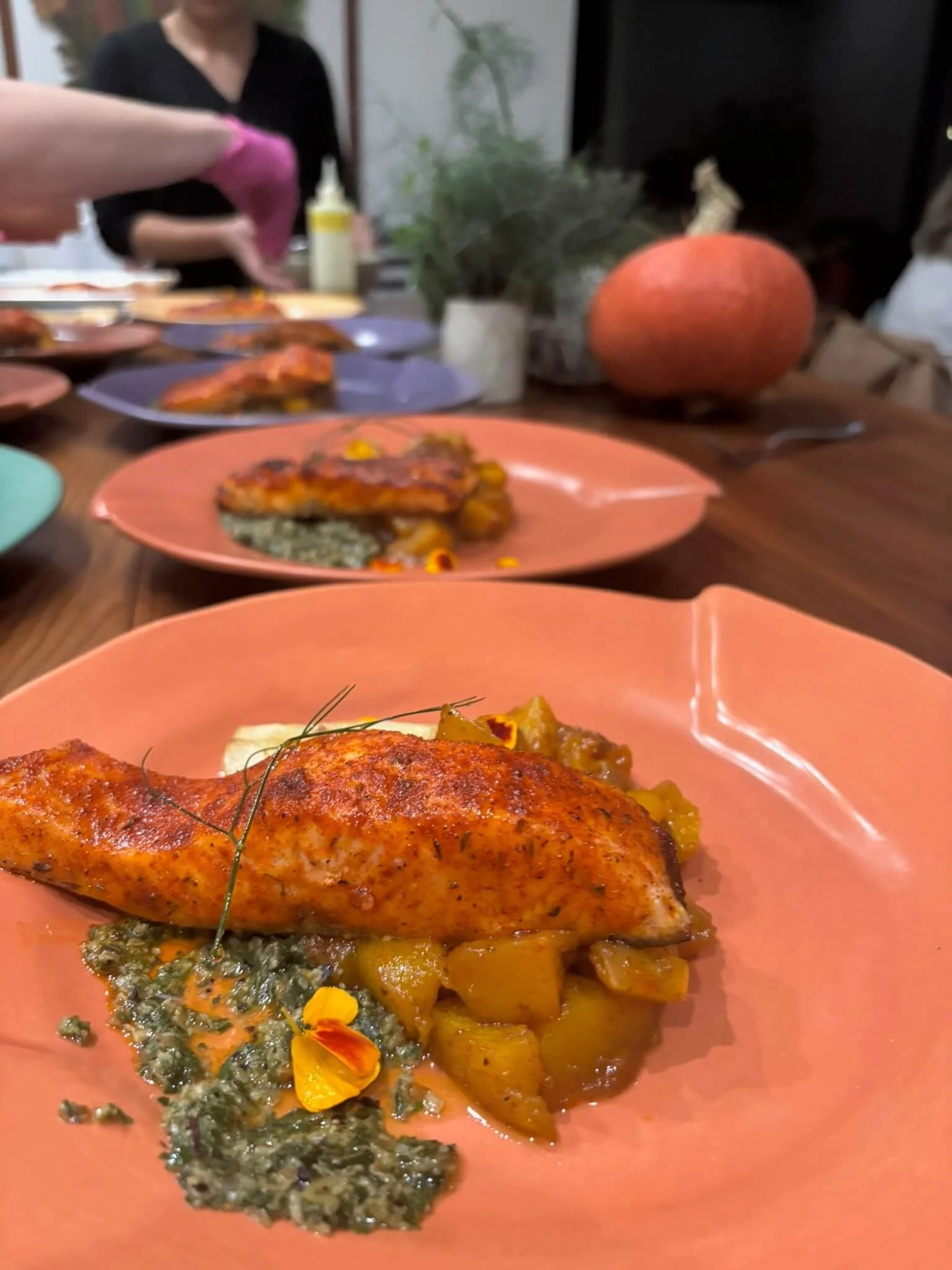 Close-up of a plated dish featuring a seasoned chicken breast, roasted vegetables, mashed potatoes topped with herbs, and garnished with edible flowers, set on a pink plate on a wooden table.