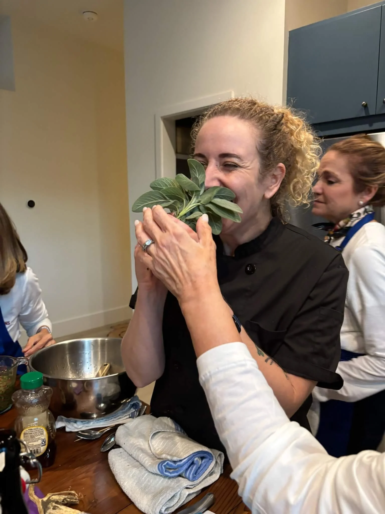A woman with curly blonde hair and a black chef jacket is smelling a bunch of fresh green herbs, held by another person with an white shirt. The scene takes place in a kitchen with a wooden table, a metal bowl, and various ingredients.