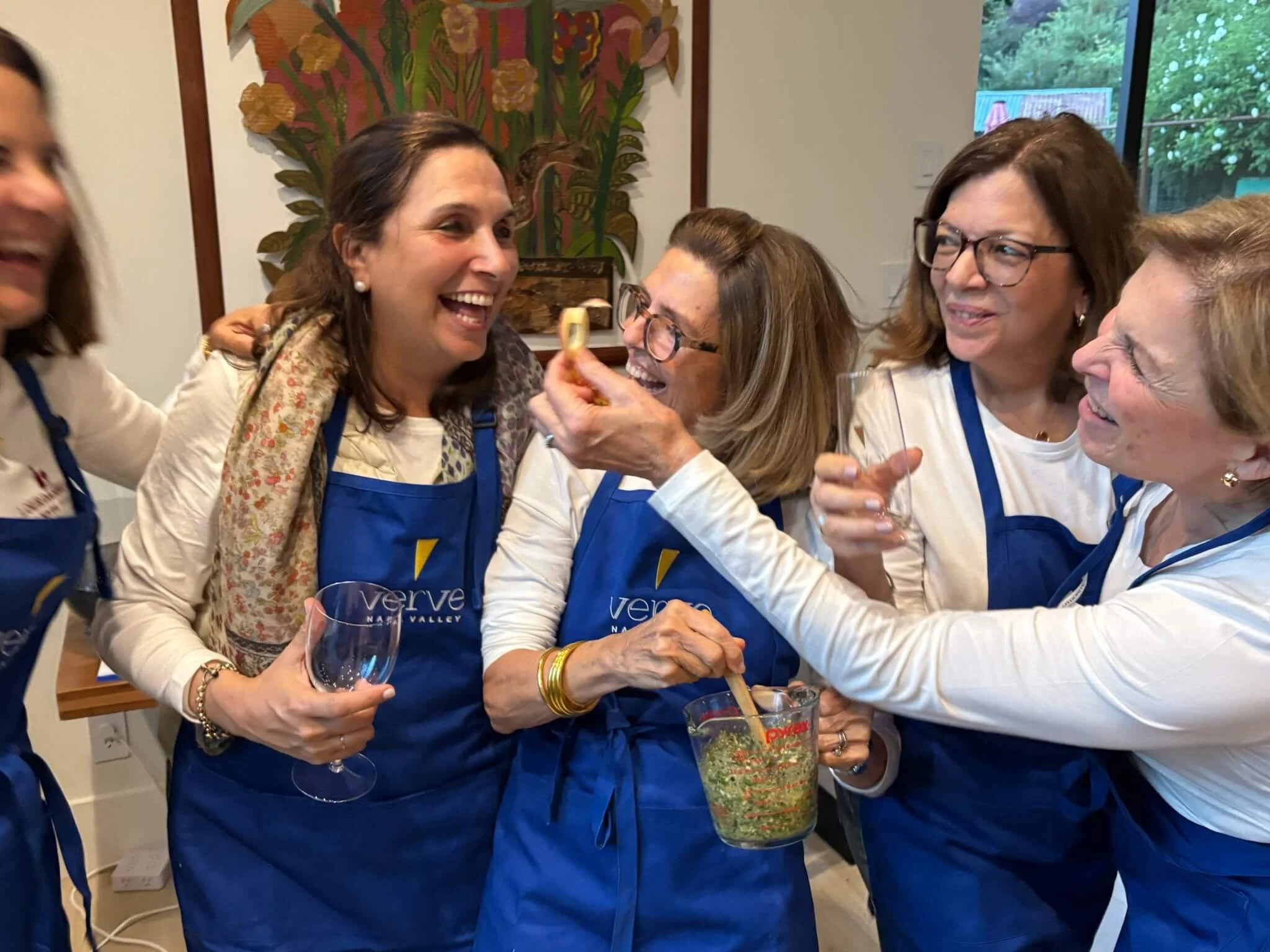Group of women wearing blue aprons, smiling and laughing during a cooking or food tasting activity, with some holding glasses and one woman stirring a bowl of food.