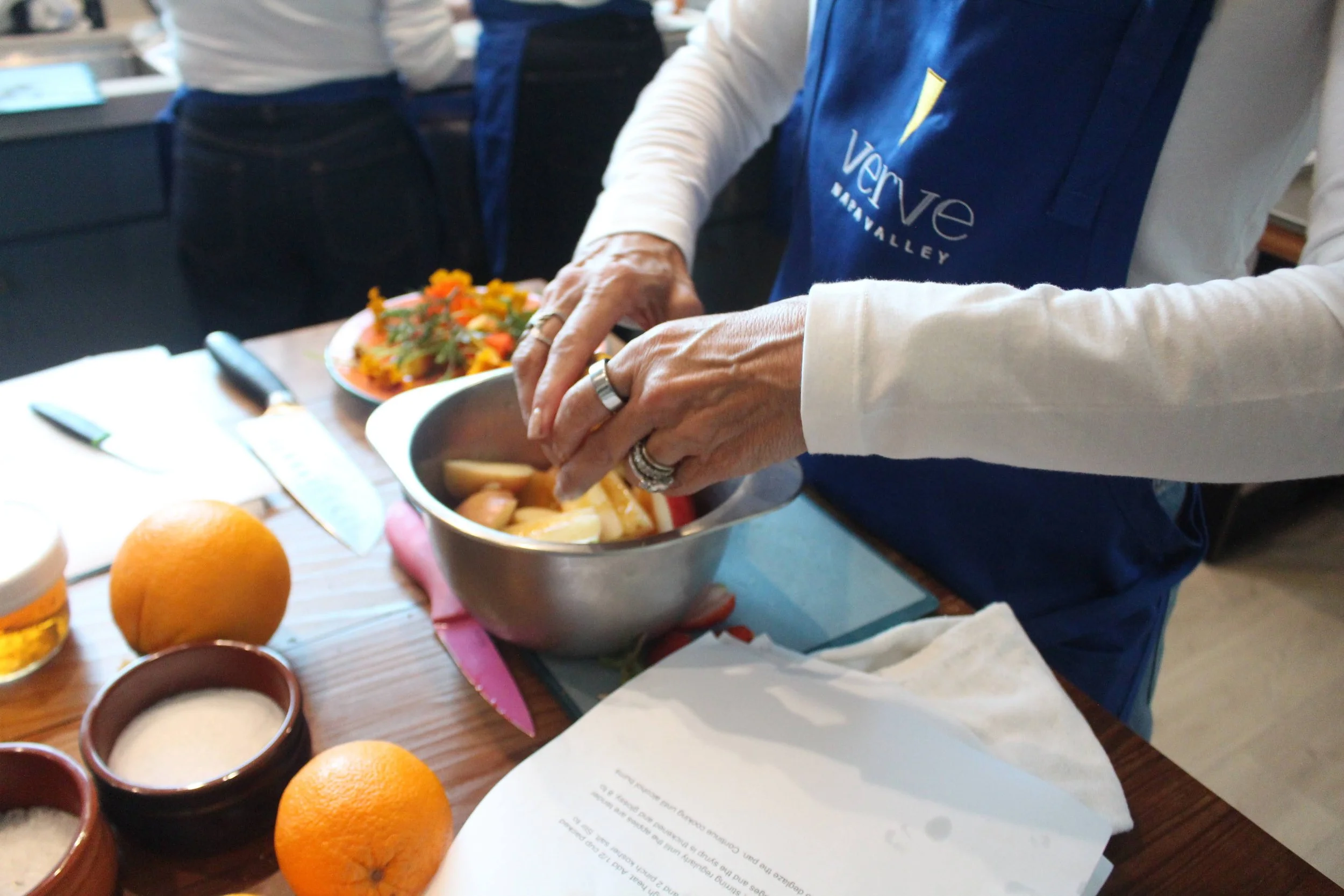 A person wearing a blue apron is preparing food in a kitchen, mixing ingredients in a metal bowl. There are oranges, a glass of beer, and a plate of salad on the counter, along with a knife and a cutting board.