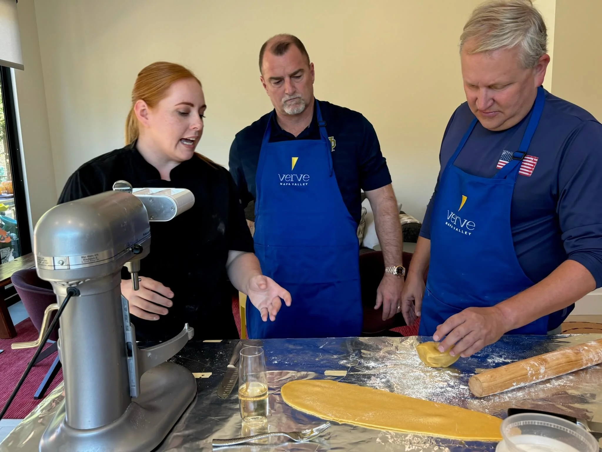 Three people, two men and one woman, wearing blue aprons with 'Verve Napa Valley' logo, making pasta in a kitchen with a pasta roller and ingredients on the counter.