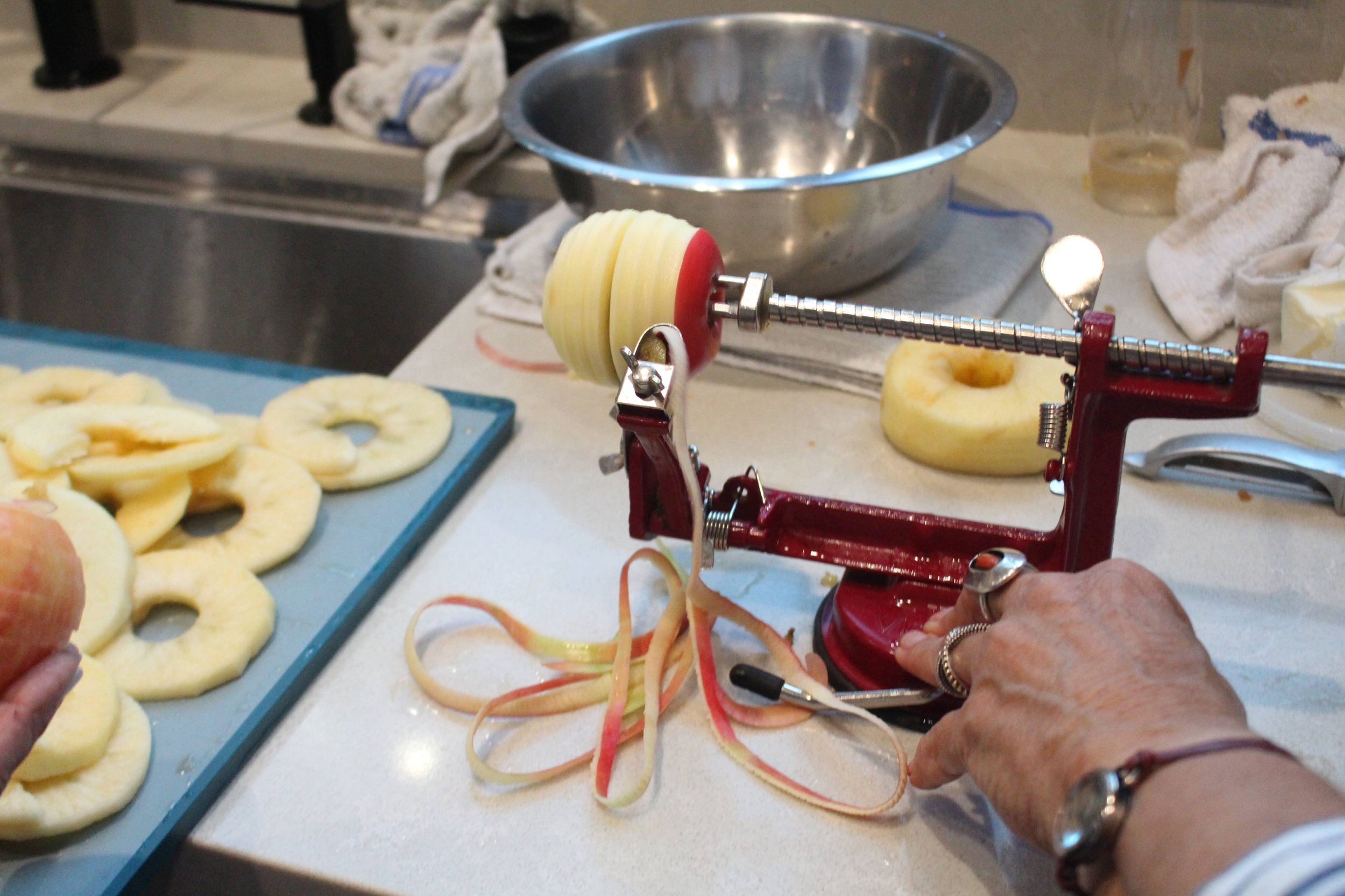 Person using a manual apple peeler machine to remove skin from an apple in a kitchen. Apple slices are on a blue tray nearby, and there are apple peels and slices on the countertop.