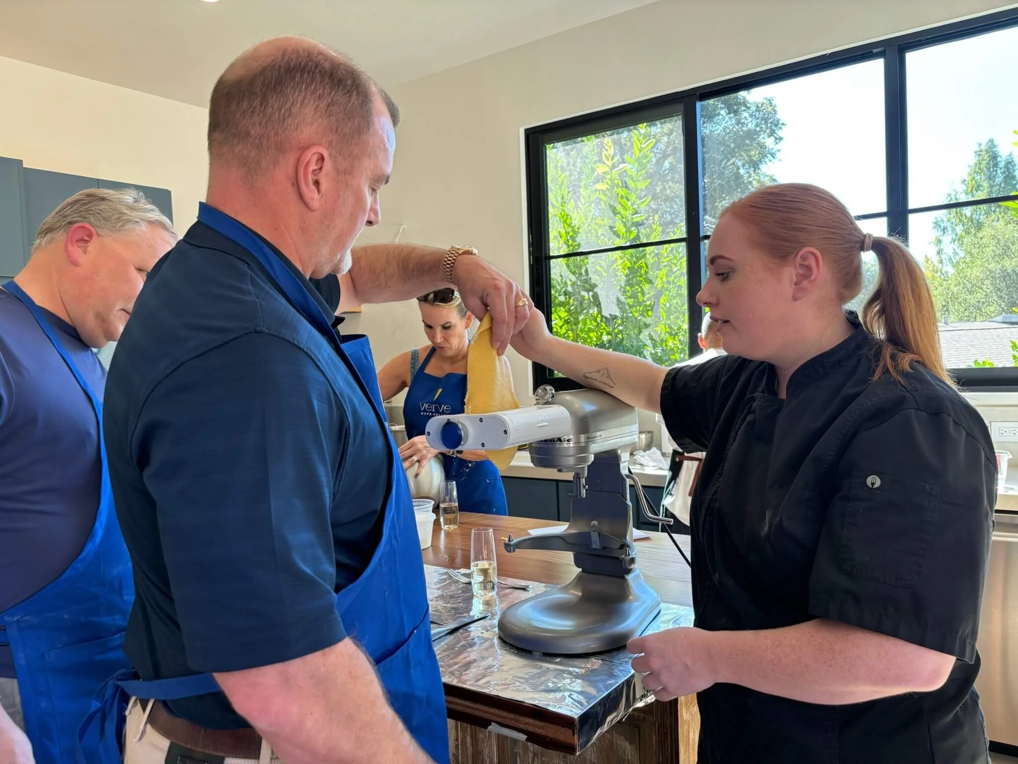 Three people in a kitchen preparing cheese using a cheese slicer, with a woman assisting and a man watching.
