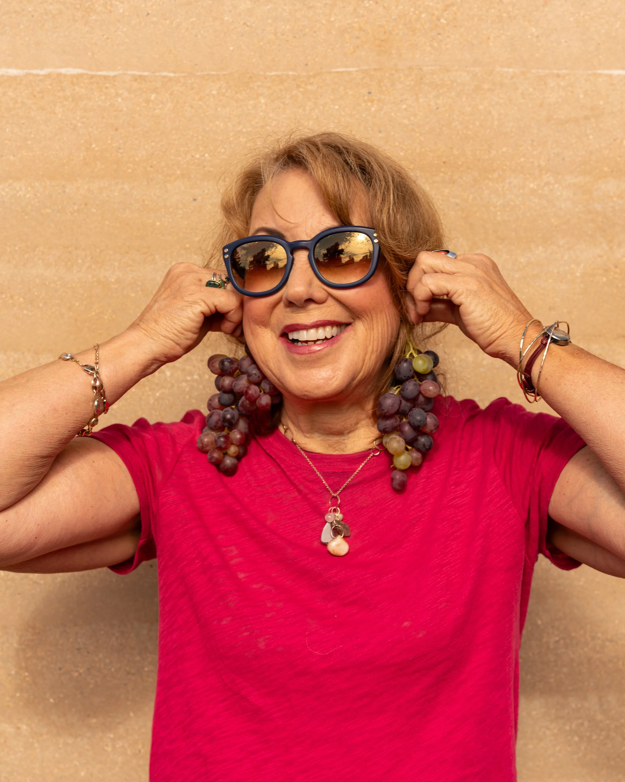 Smiling woman with grapes earrings, wearing sunglasses, jewelry, and a red shirt, standing against a beige wall, holding her sunglasses.