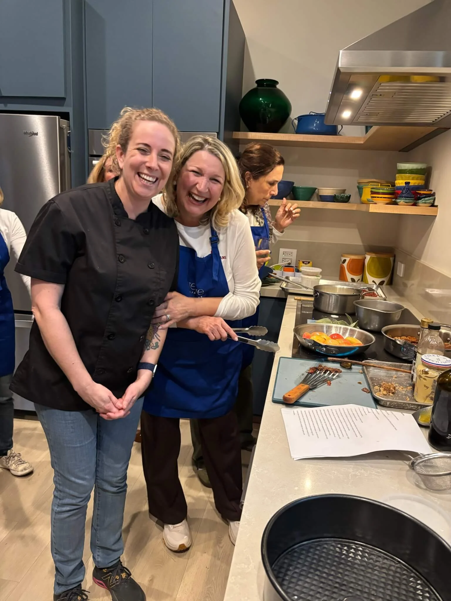 Two women smiling, one in a black chef's coat and the other in a blue apron, standing in a kitchen with another woman in the background preparing food.