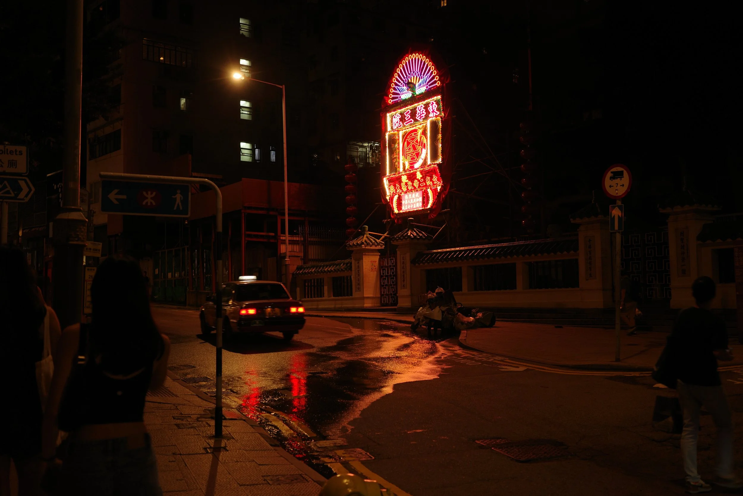 Night scene of a city street with neon signs, a car, and a group of pedestrians, with reflections on a wet road.