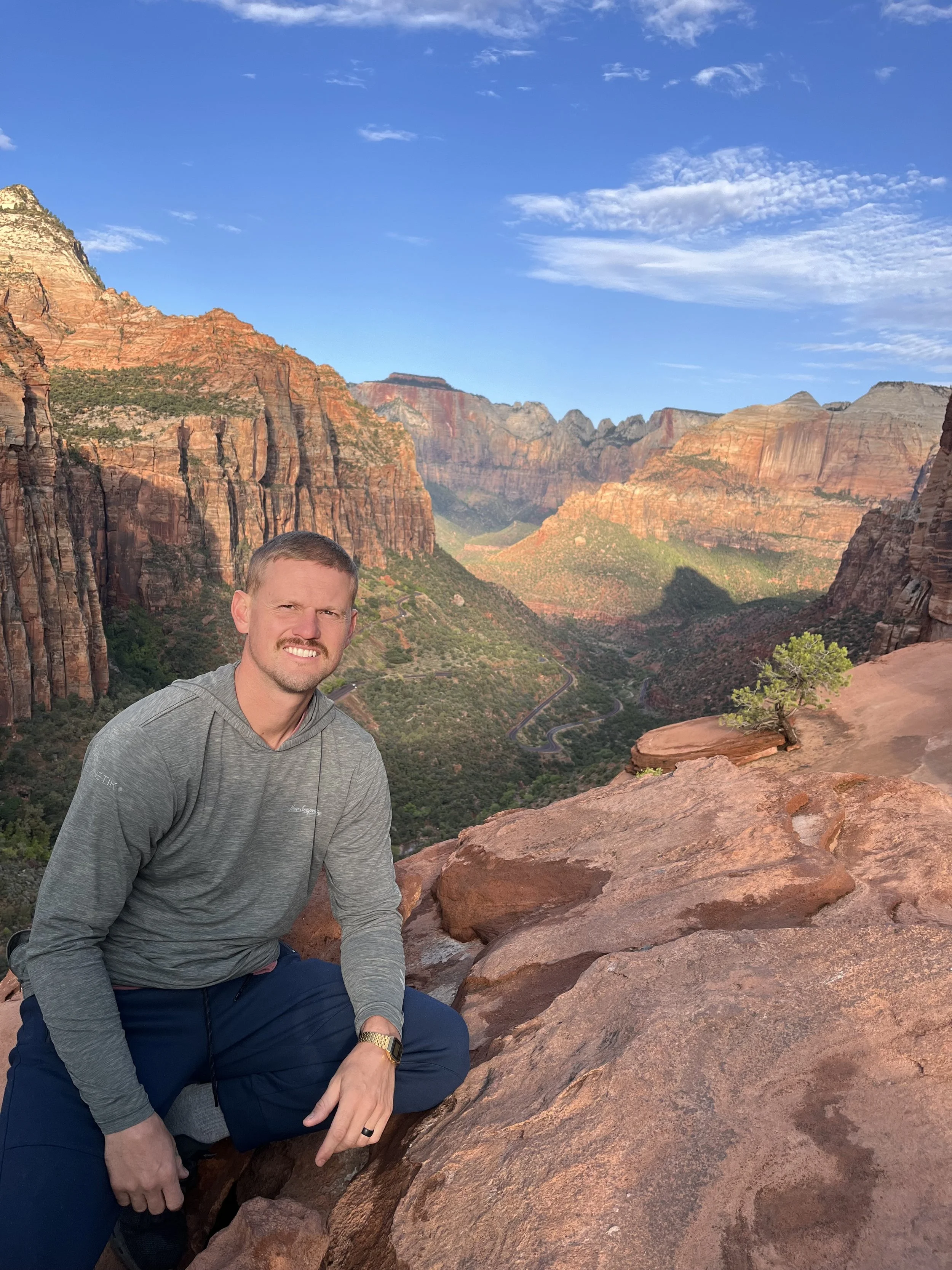 A young man with short blonde hair, wearing a light gray button-up shirt with rolled-up sleeves, standing outdoors in a grassy area with trees and a cloudy sky in the background, smiling at the camera.