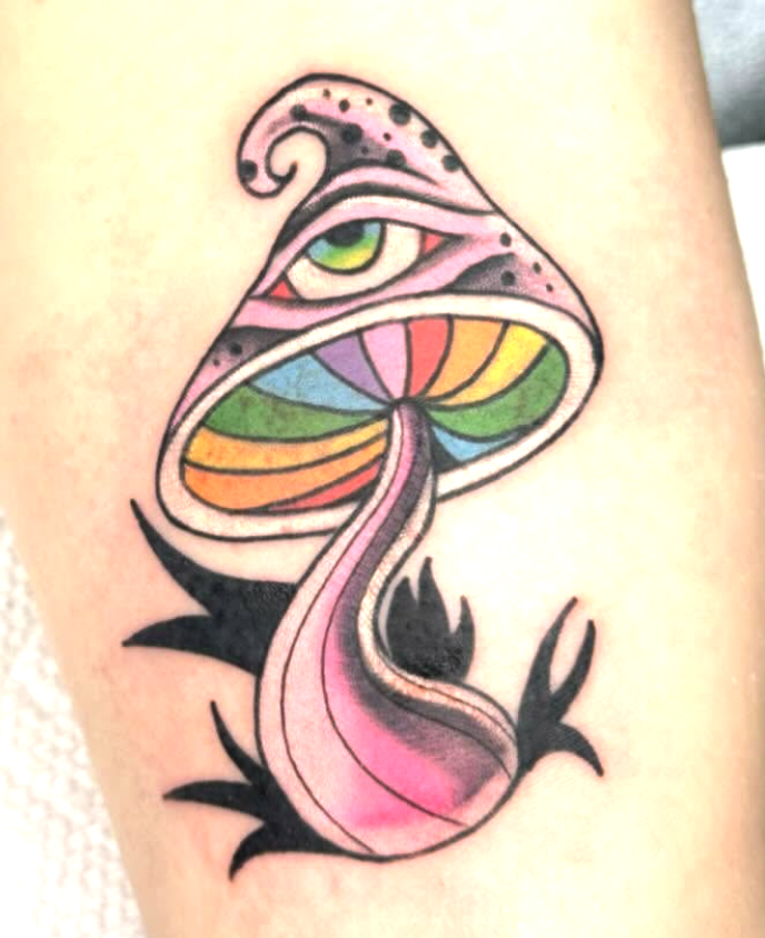 A tattoo of a mushroom with an eye on top, rainbow-colored gills, pink stem, and black leaves at the base.